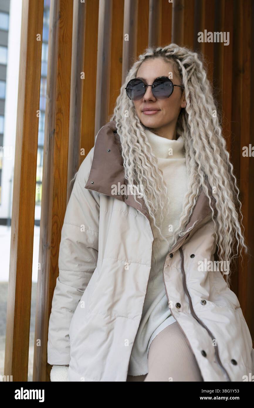 Eine Frau mit langen blonden Haaren wird mit einem weißen Mantel gesehen. Sie steht selbstbewusst und zeigt ihre elegante Kleidung mit Pose und Anmut Stockfoto