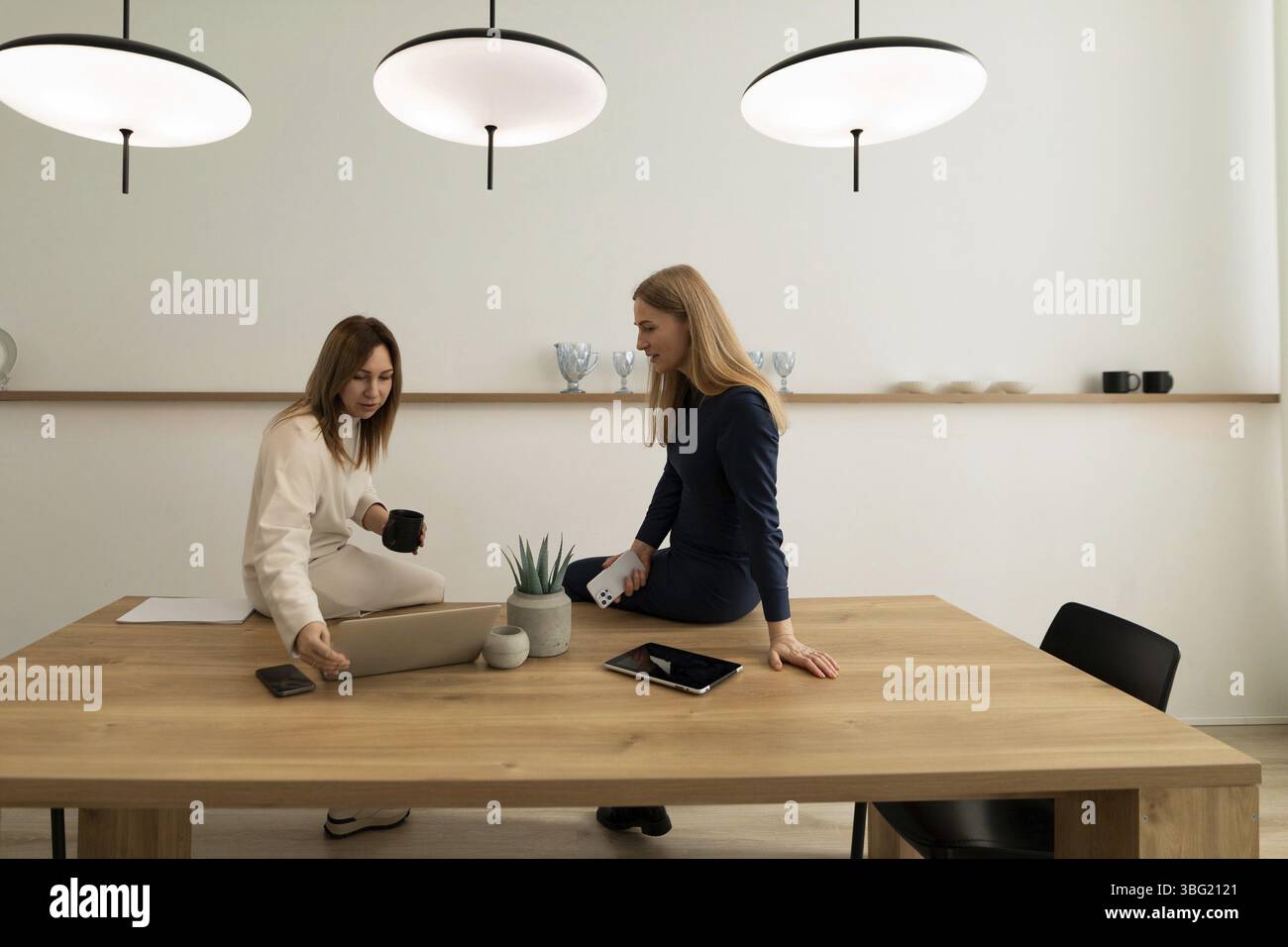 Zwei Frauen führen produktive Gespräche, eine sitzt auf dem Tisch und die andere lehnt sich über einen Laptop in einem stilvollen und gut beleuchteten Raum Stockfoto