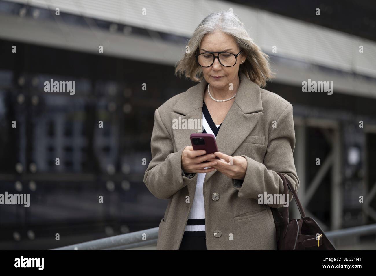 Eine ältere Geschäftsfrau in einem schicken Outfit geht draußen und konzentriert sich auf ihr Smartphone. Der Hintergrund zeigt moderne Architektur, die hervorhebt Stockfoto