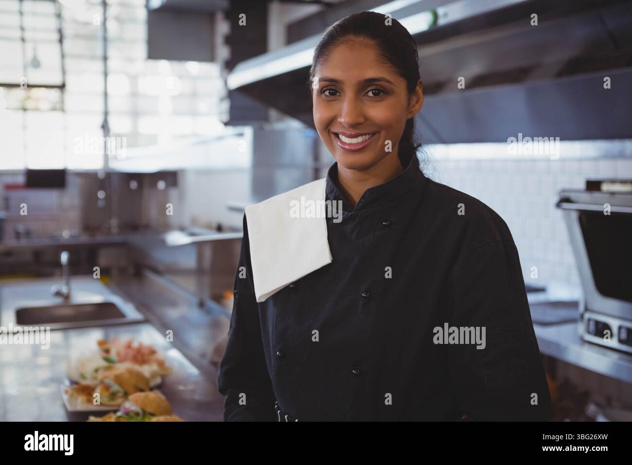 Indische Küchenchefin in schwarzer Jacke lächelt in der Küche neben der Zubereitungstheke mit belegten Sandwiches Stockfoto