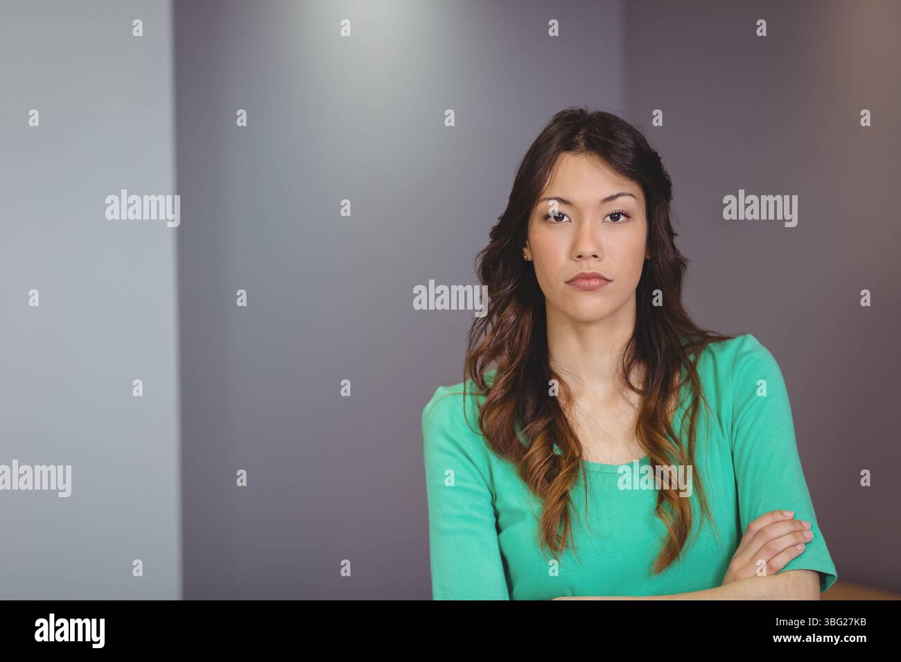 Frau mit grünem, langärmeligem Oberteil stehend mit gekreuzten Armen im Studio an grauer Wand, Kopierraum Stockfoto