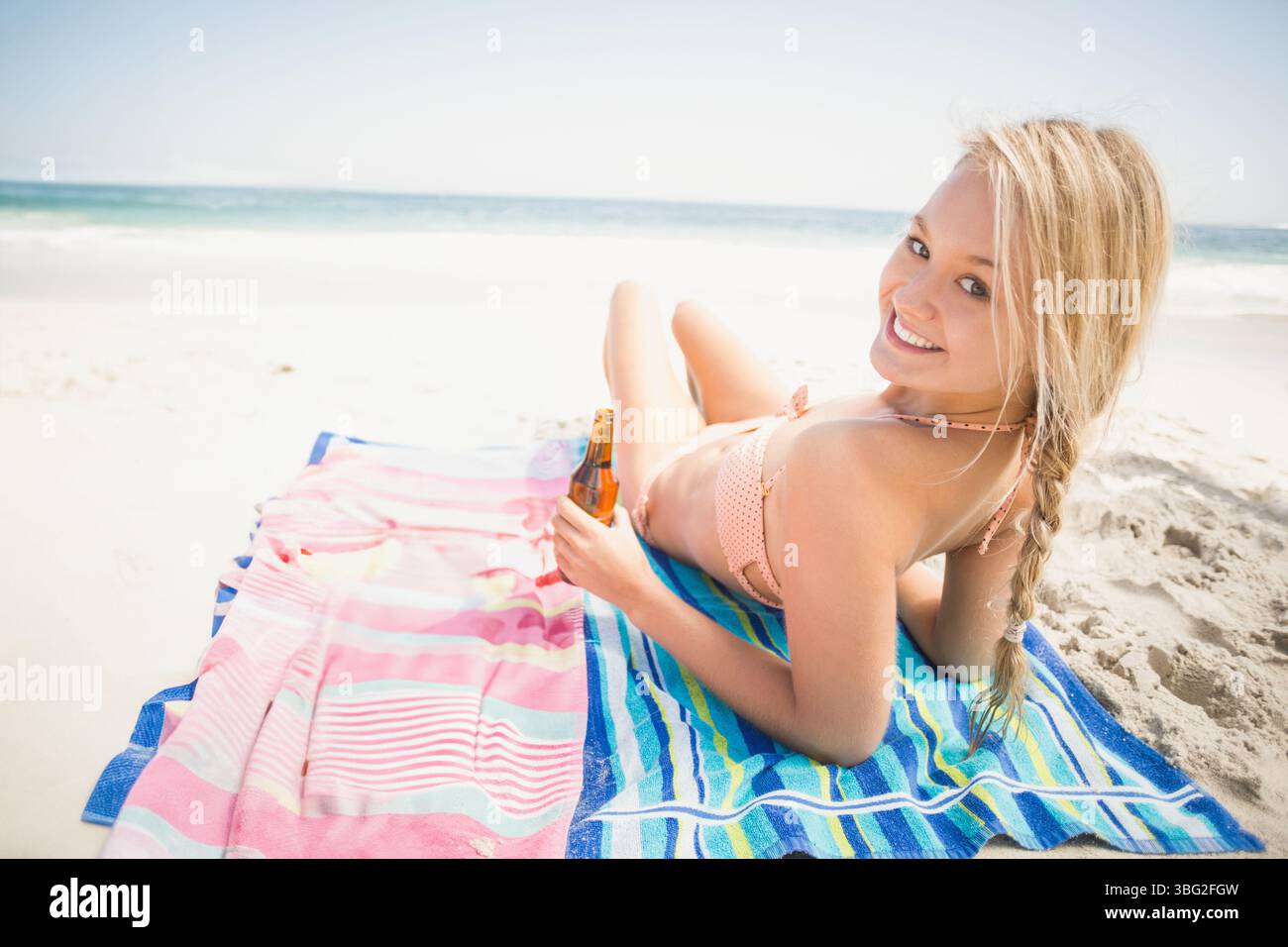 Frau, die auf einem gestreiften Handtuch am Sandstrand liegt, in rosa Bikinizone, mit brauner Flasche, Kopierraum Stockfoto