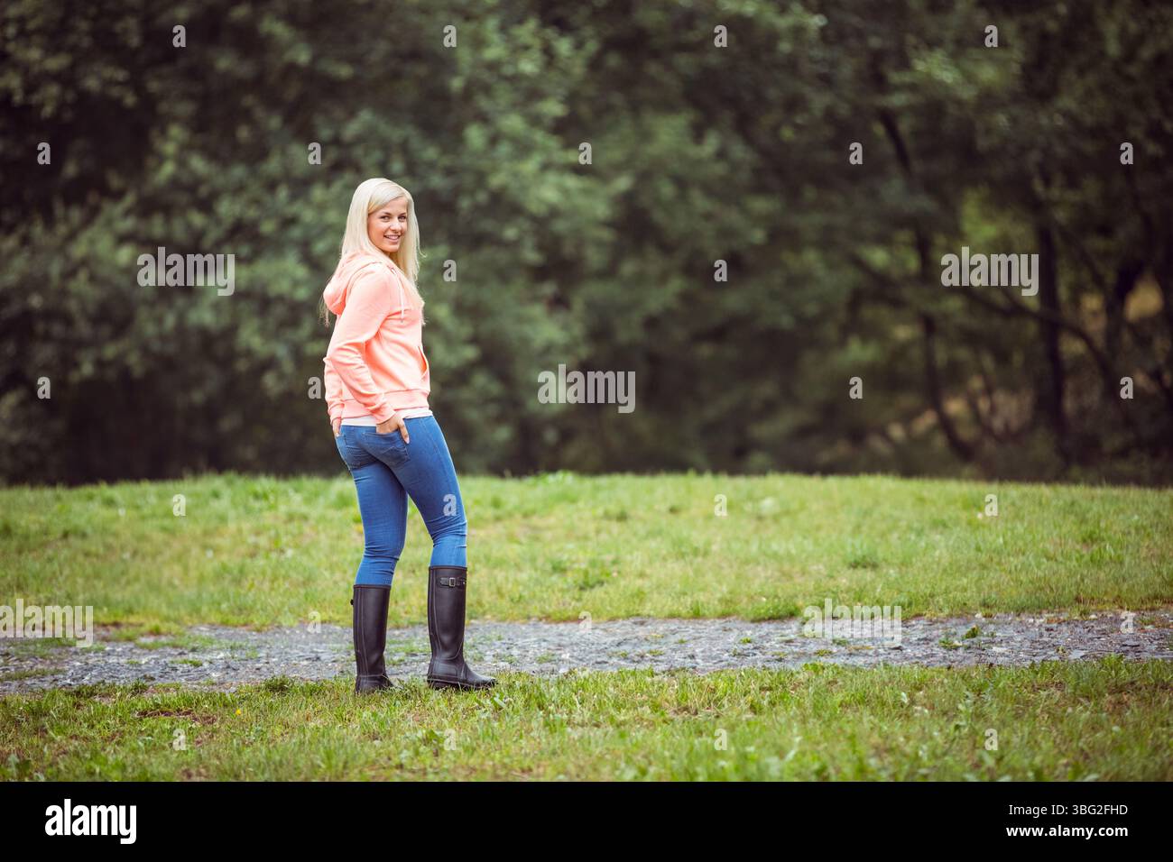 Frau mit rosafarbenem Hoodie, Jeans, schwarzen Gummistiefeln, die auf einem Kiesweg zwischen Gras in der Nähe von Bäumen stehen Stockfoto