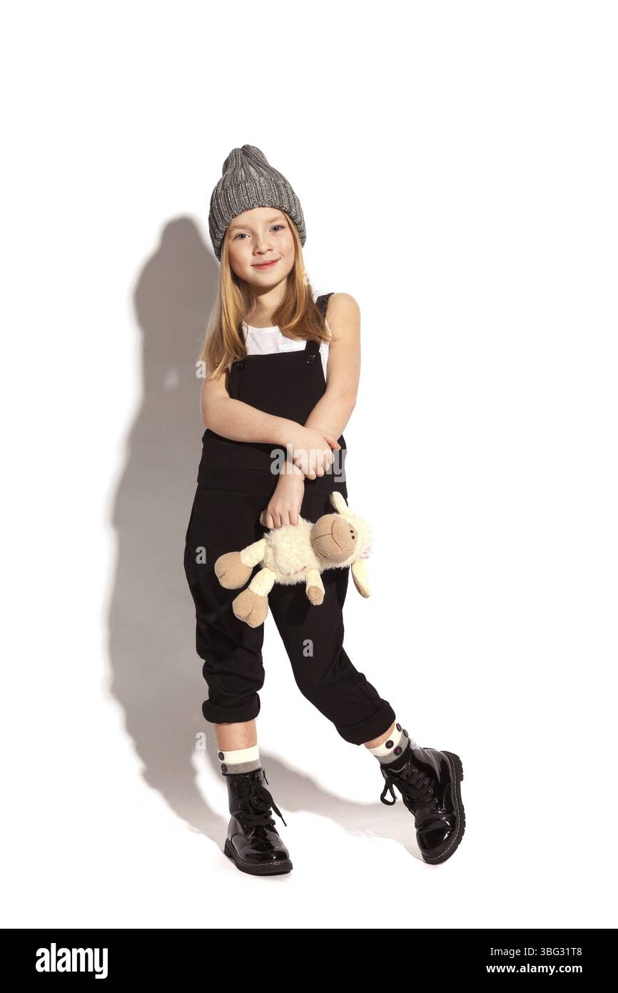 Isoliert auf weißem, lächelndem rothaarigem Mädchen in schwarzem Overall, weißem T-Shirt, grauem Hut und lackierten Schnürschuhen, die ein Plüschtier halten. Copyspace Stockfoto