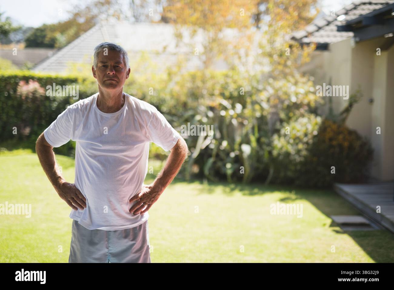 Senior Mann, der mit Händen auf Hüften auf dem Garten in der Nähe der Hecke unter hellem Sonnenlicht steht Stockfoto