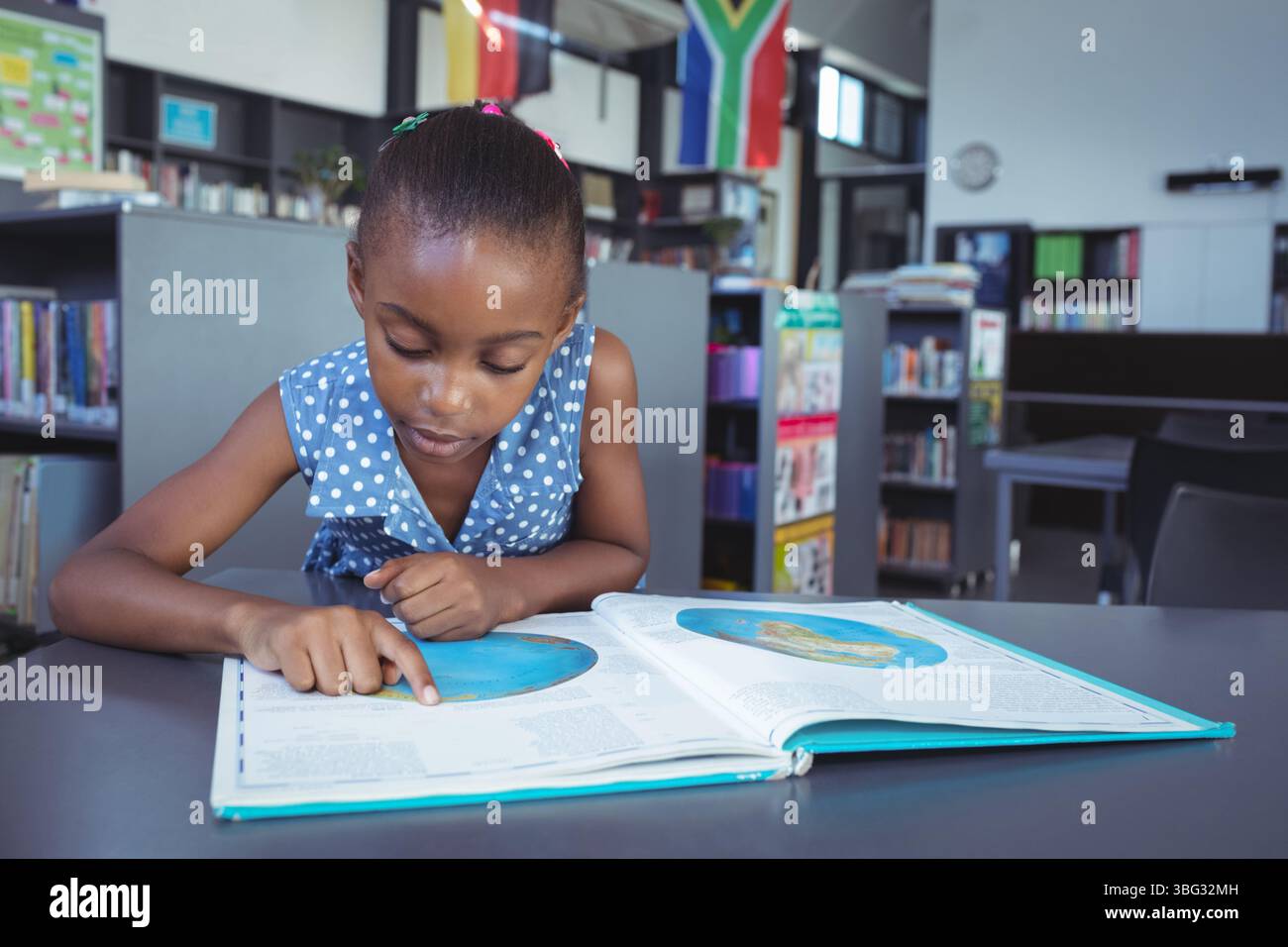 Afroamerikanisches Schulmädchen, das sich über den offenen atlas in der öffentlichen Bibliothek beugt und auf die Karte zeigt Stockfoto