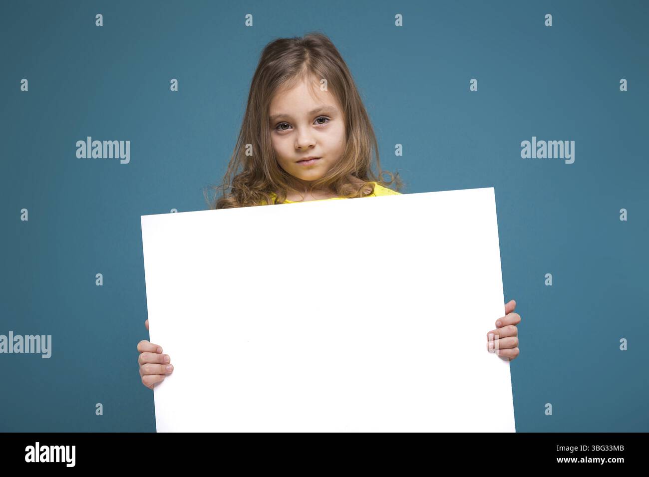 Isoliert auf blauem, wunderschönem kaukasischem Kind mit langen Haaren, in gelbem T-Shirt und schwarzer Hose halten großes leeres Poster, gedreht, Blick nach unten Stockfoto