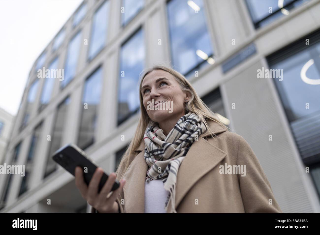 Eine Frau mit langem Mantel und Schal läuft an einem hohen, modernen Gebäude mit vielen großen Fenstern vorbei. Sie schaut hoch und hält einen schwarzen Smartph Stockfoto