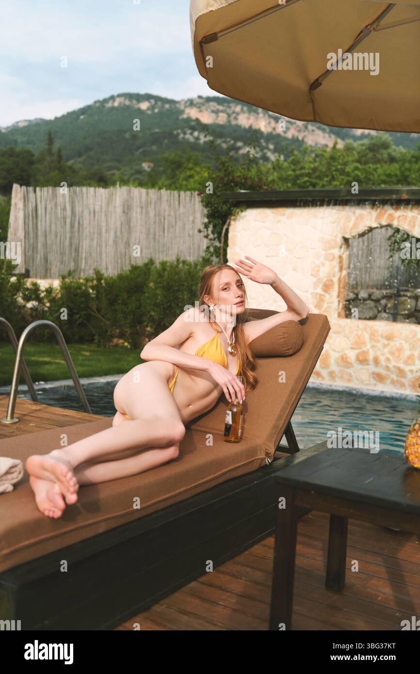Eine blonde Frau aus den 20er Jahren liegt auf einer Liege am privaten Pool und genießt in Ruhe ein erfrischendes Getränk. Stockfoto