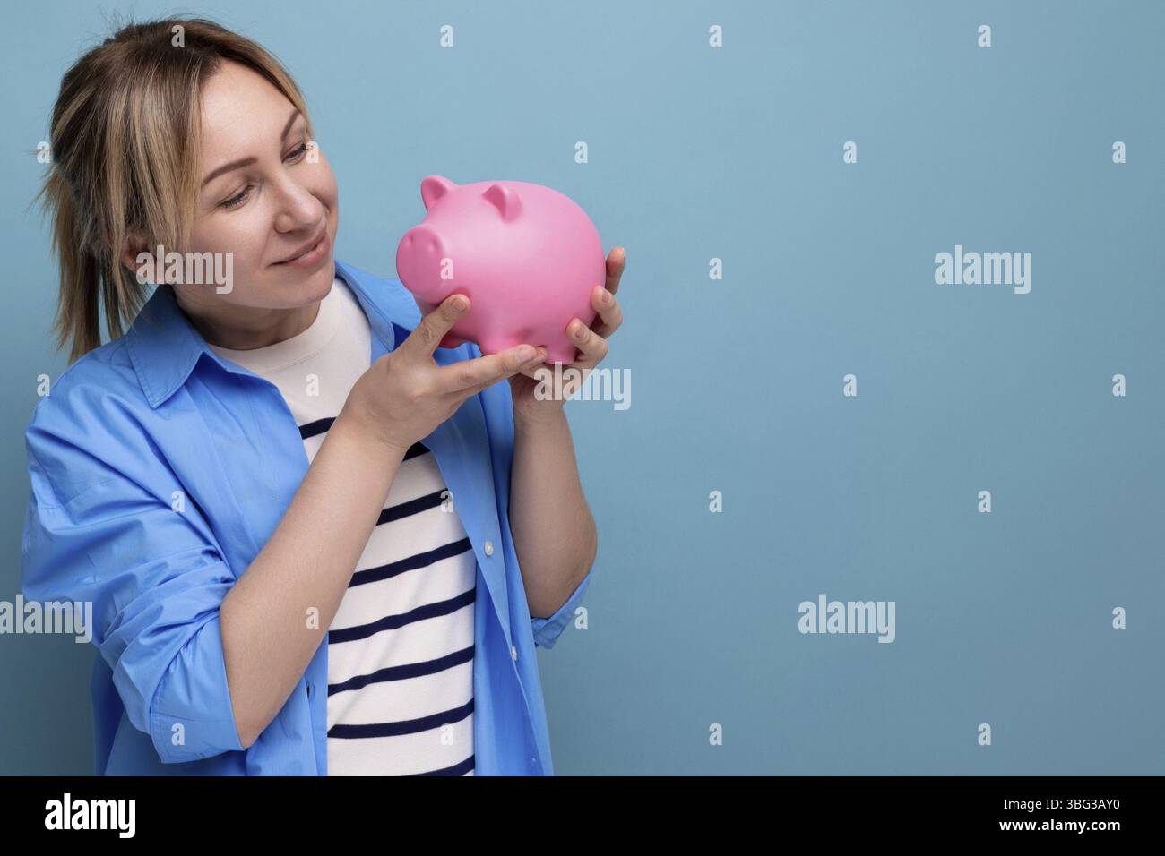 Blonde attraktive junge Frau in lässiger Kleidung, die nachdenklich eine Sparkasse mit Geld auf blauem Hintergrund mit Kopierraum hält Stockfoto