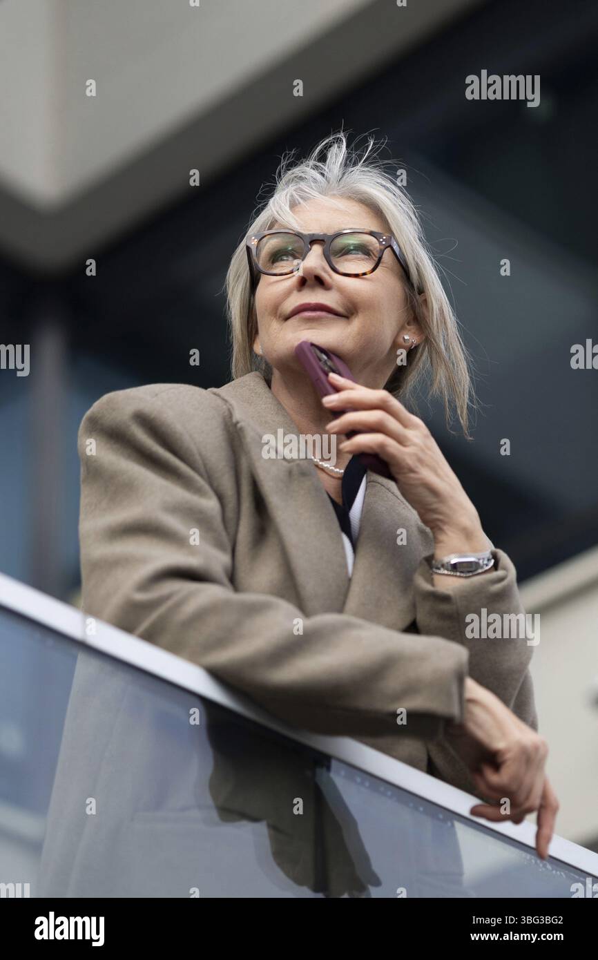 Eine ältere Frau steht entspannt auf einem Balkon, in Gedanken verloren, während sie ihr Handy nahe am Kinn hält, und genießt einen Moment der Besinnung in einem kontempo Stockfoto