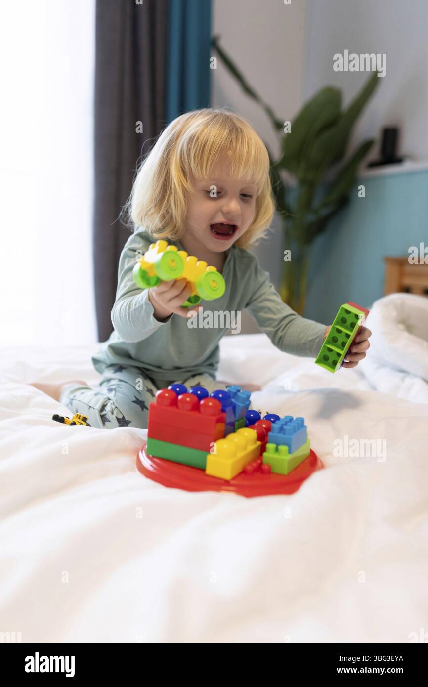 Ein Kleinkind baut gerne ein farbenfrohes Gebäude mit Bausteinen, während es auf einem bequemen Bett sitzt. Natürliches Licht erhellt fröhliche bei Stockfoto