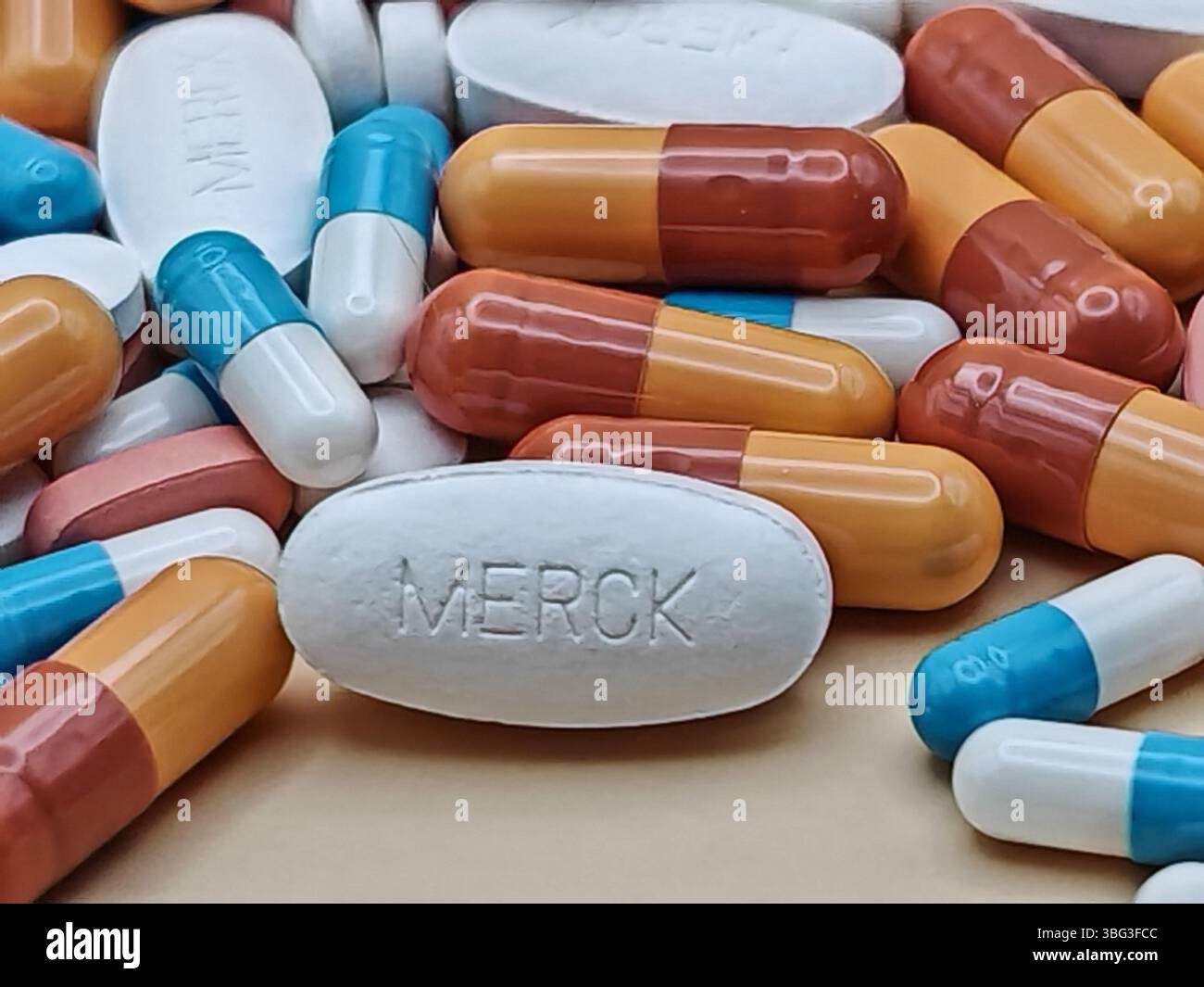 Weiße Pille mit MERCK-Aufdruck auf Tablettenstapel und farbenfrohe Kapseln in pharmazeutischer Optik. Stockfoto