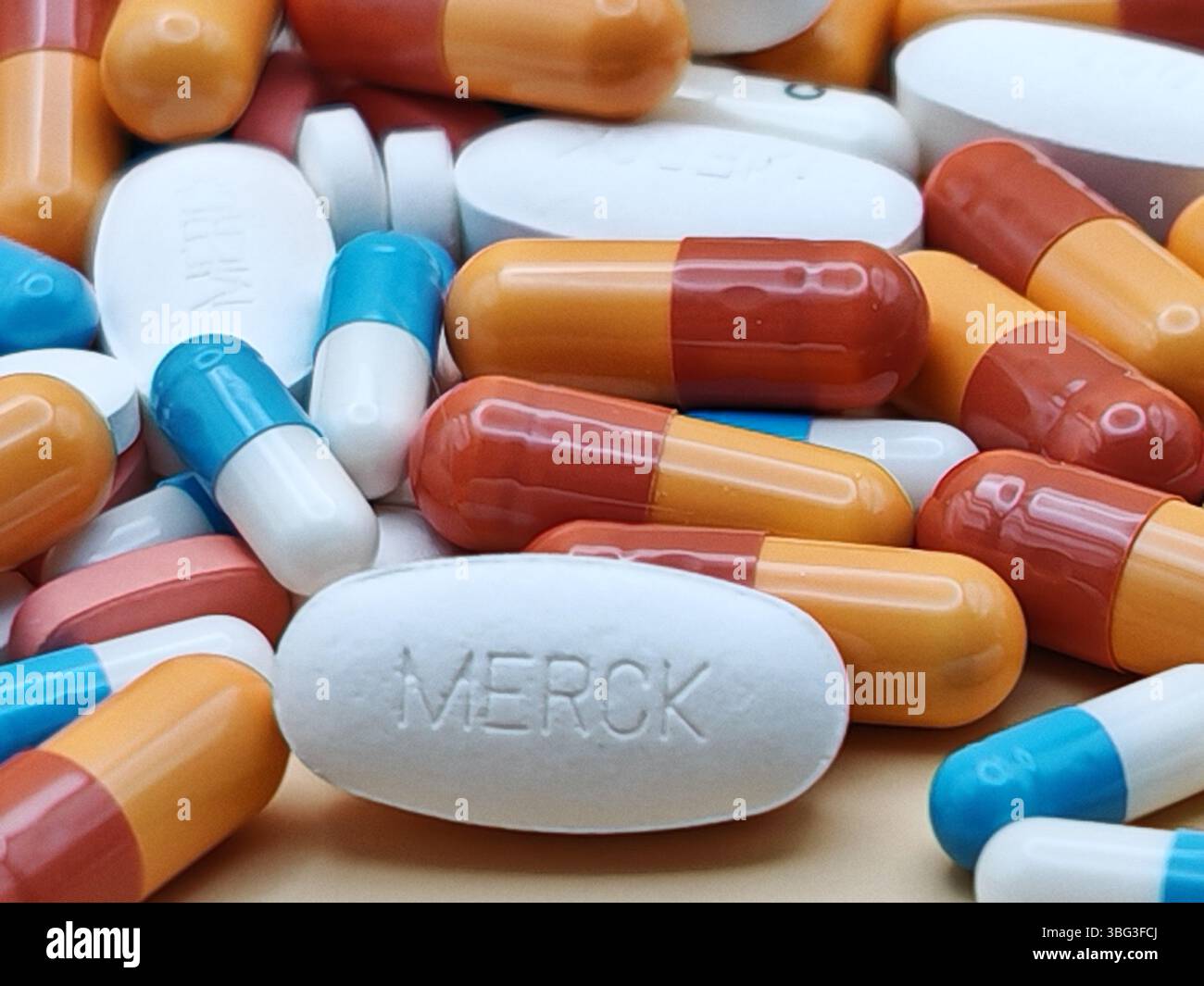 Weiße Pille mit MERCK-Aufdruck auf Tablettenstapel und farbenfrohe Kapseln in pharmazeutischer Optik. Stockfoto