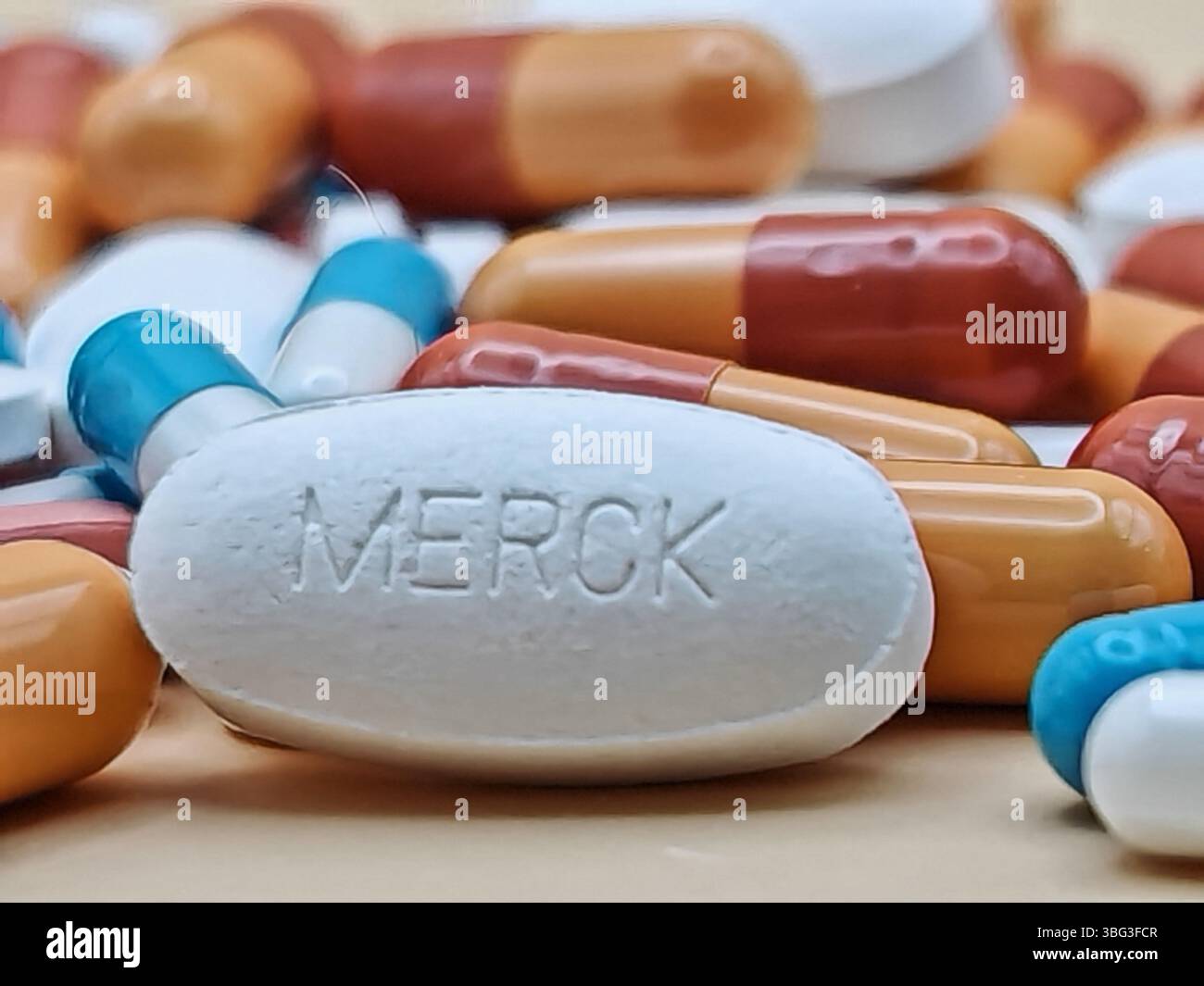 Weiße Pille mit MERCK-Aufdruck auf Tablettenstapel und farbenfrohe Kapseln in pharmazeutischer Optik. Stockfoto