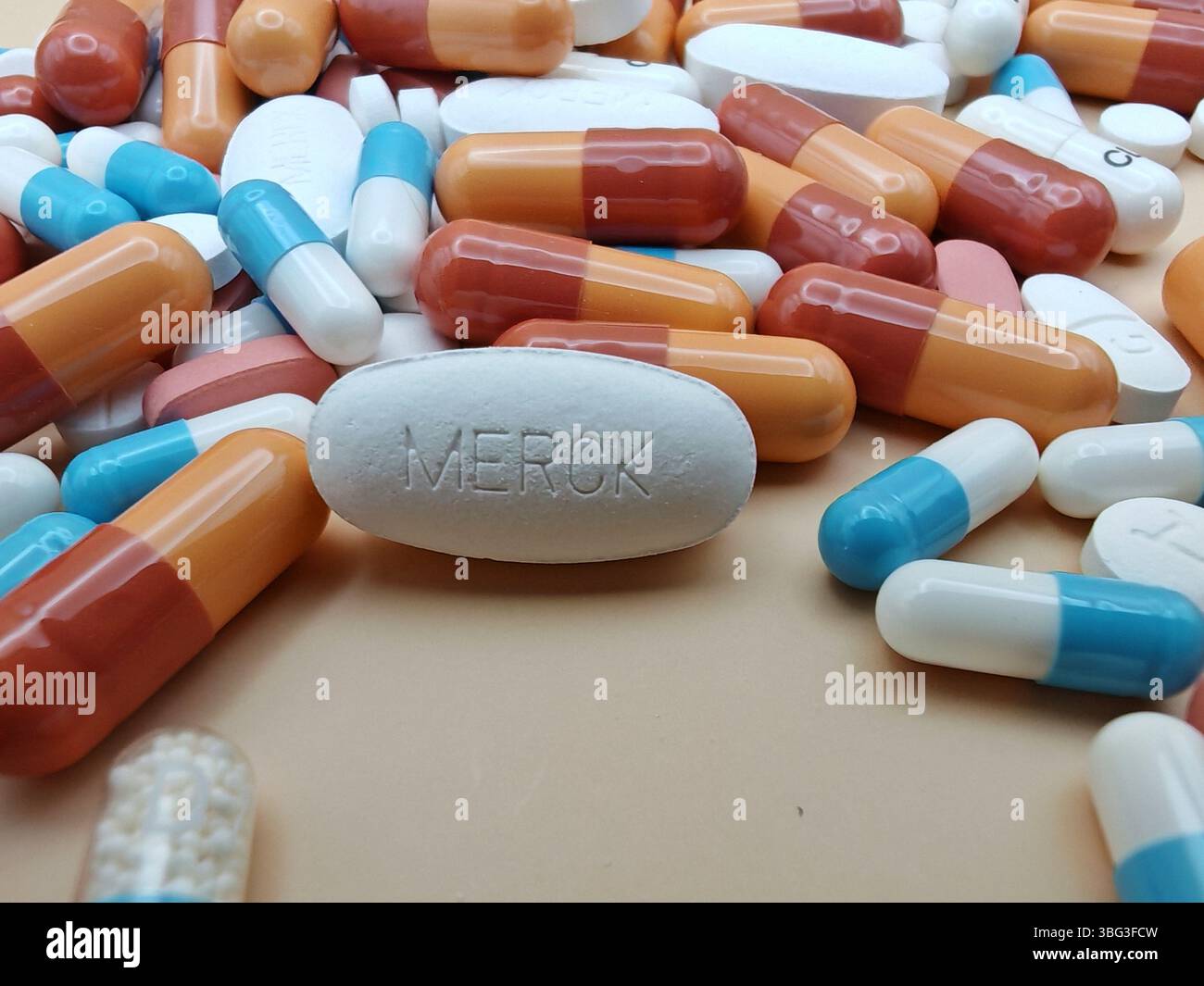 Weiße Pille mit MERCK-Aufdruck auf Tablettenstapel und farbenfrohe Kapseln in pharmazeutischer Optik. Stockfoto