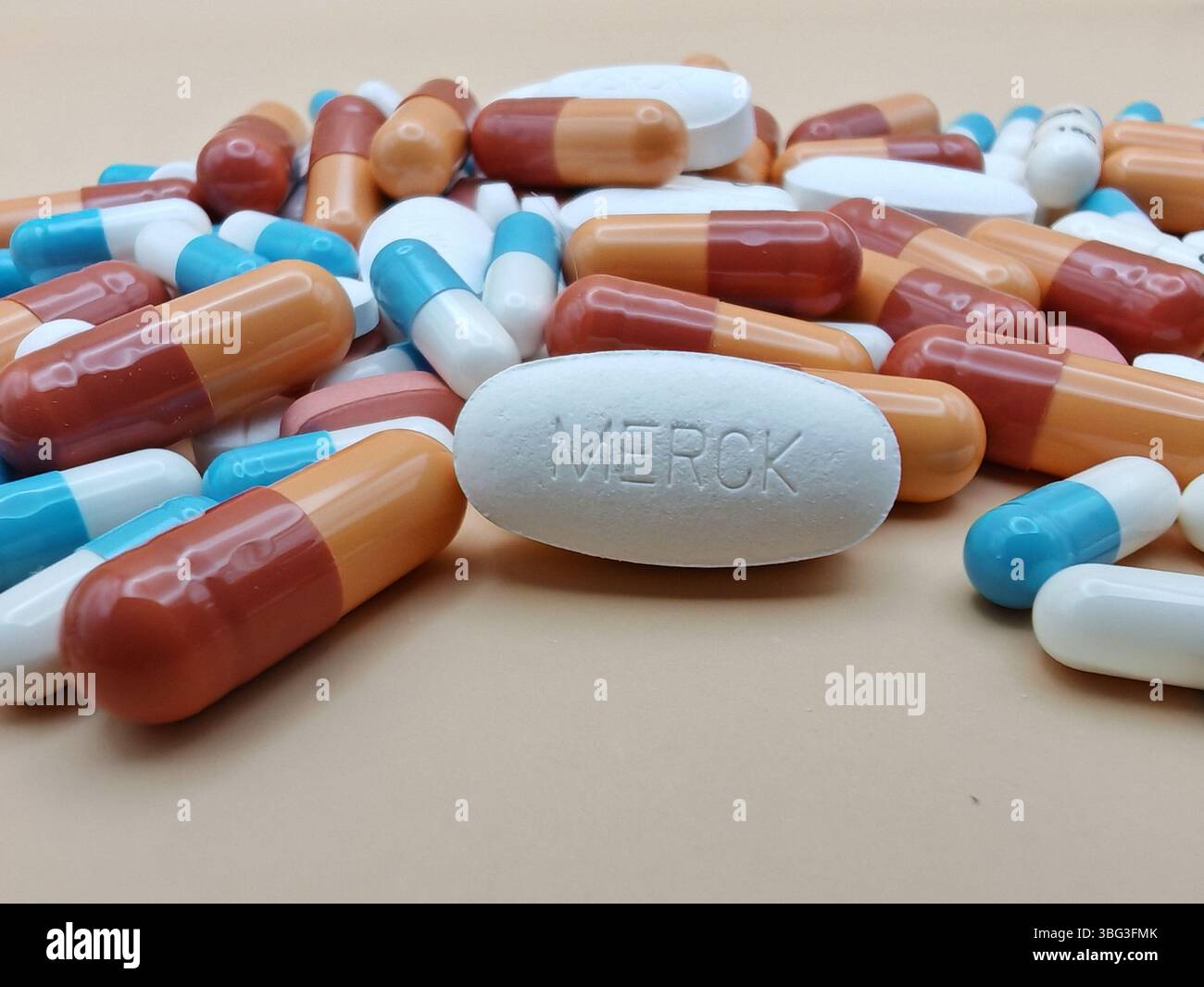 Weiße Pille mit MERCK-Aufdruck auf Tablettenstapel und farbenfrohe Kapseln in pharmazeutischer Optik. Stockfoto