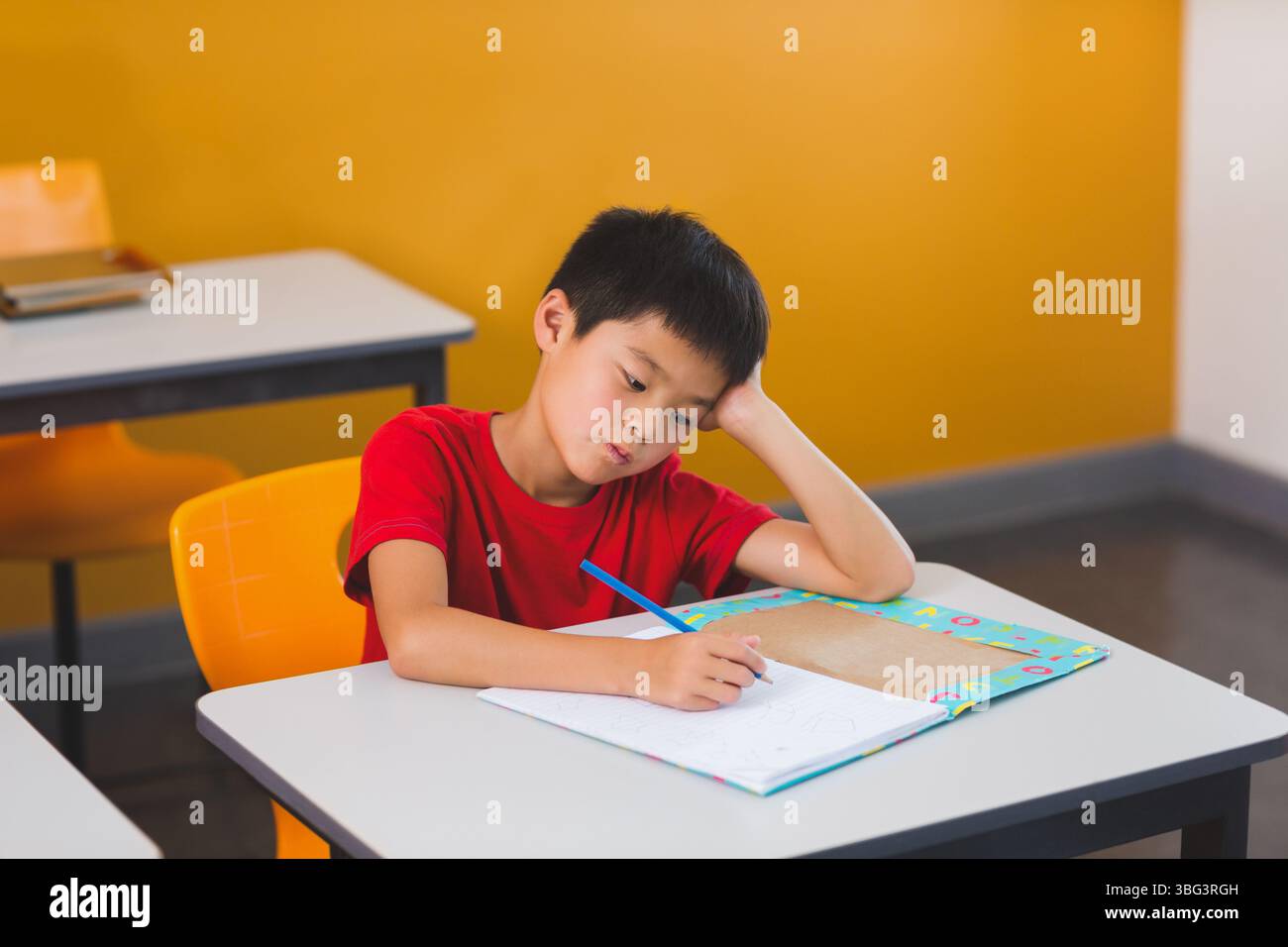 Asiatischer Junge, der Kopf auf der Hand liegt und in einem bunten Arbeitsbuch am Schulpult mit Bleistift schreibt Stockfoto