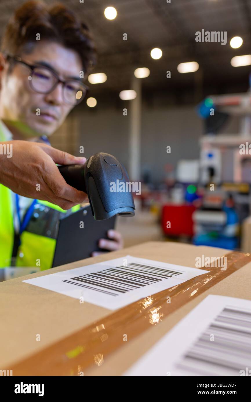 Asiatischer Mann in Sicherheitsweste, der Barcode auf der Kiste scannt und die Zwischenablage im Lager hält, im Kopierraum Stockfoto