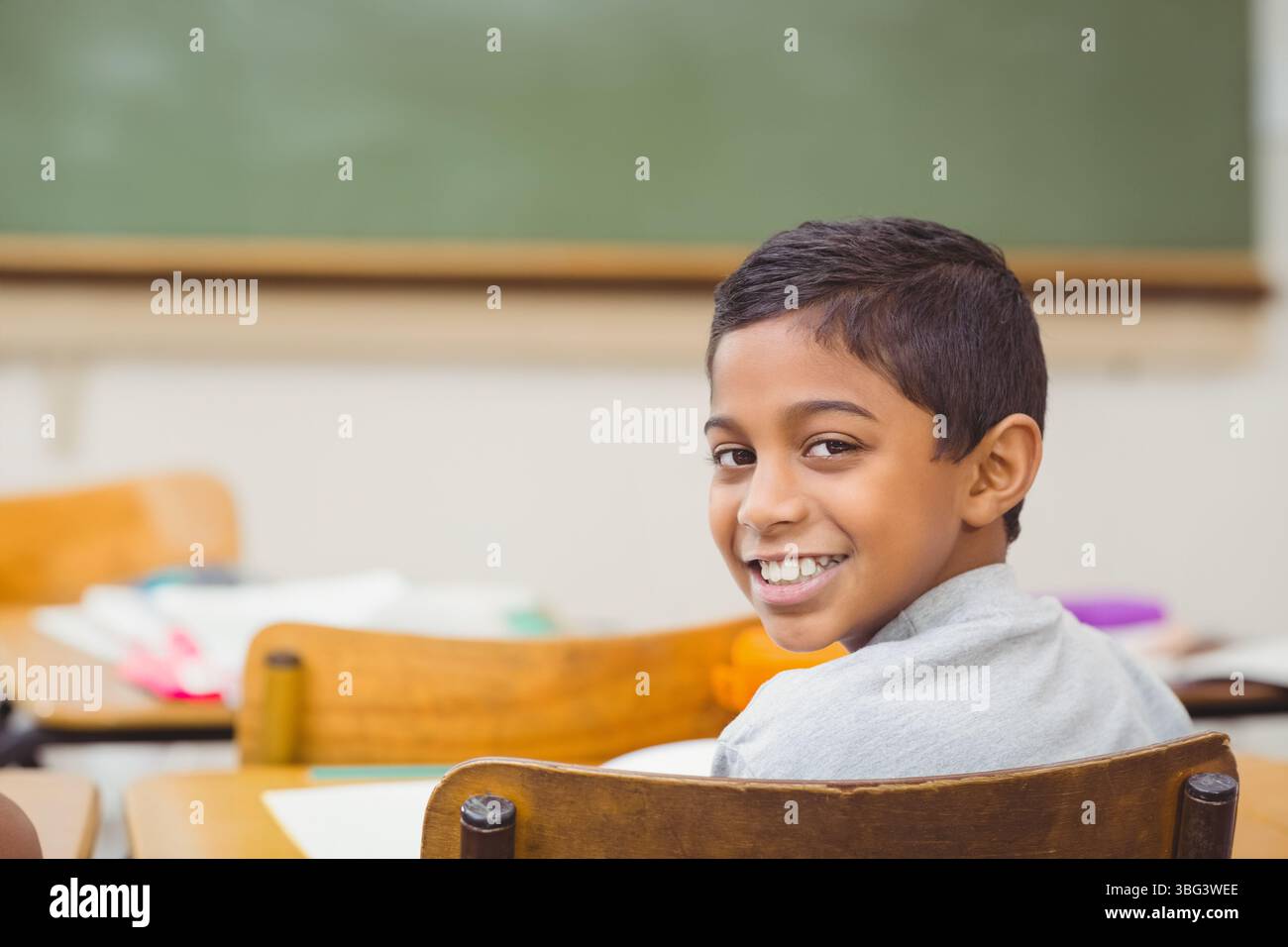 Asiatischer Junge, der am Holzschreibtisch sitzt, im Klassenzimmer auf Papieren unter Tafel schreibt, Kopierraum Stockfoto