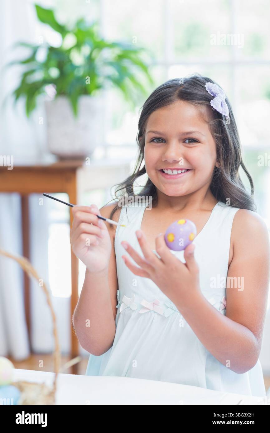 Schulmädchen malt Ei am Tisch am hellen Fenster zu Hause mit Korb und Pinsel Stockfoto