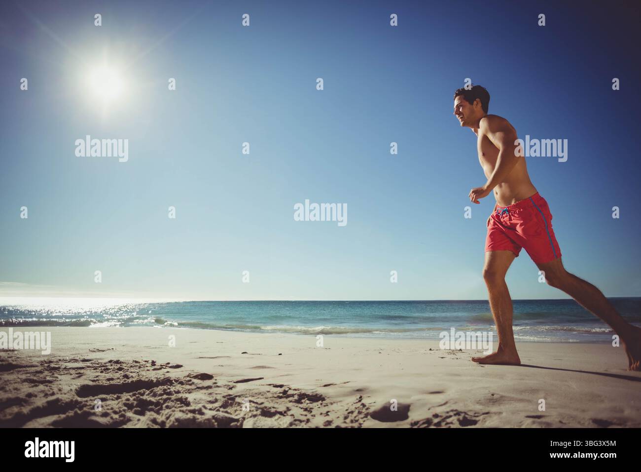 Läufer ohne Hemd, Joggen auf nassem Sand unter strahlender Sonne mit Meereswellen, Kopierraum Stockfoto