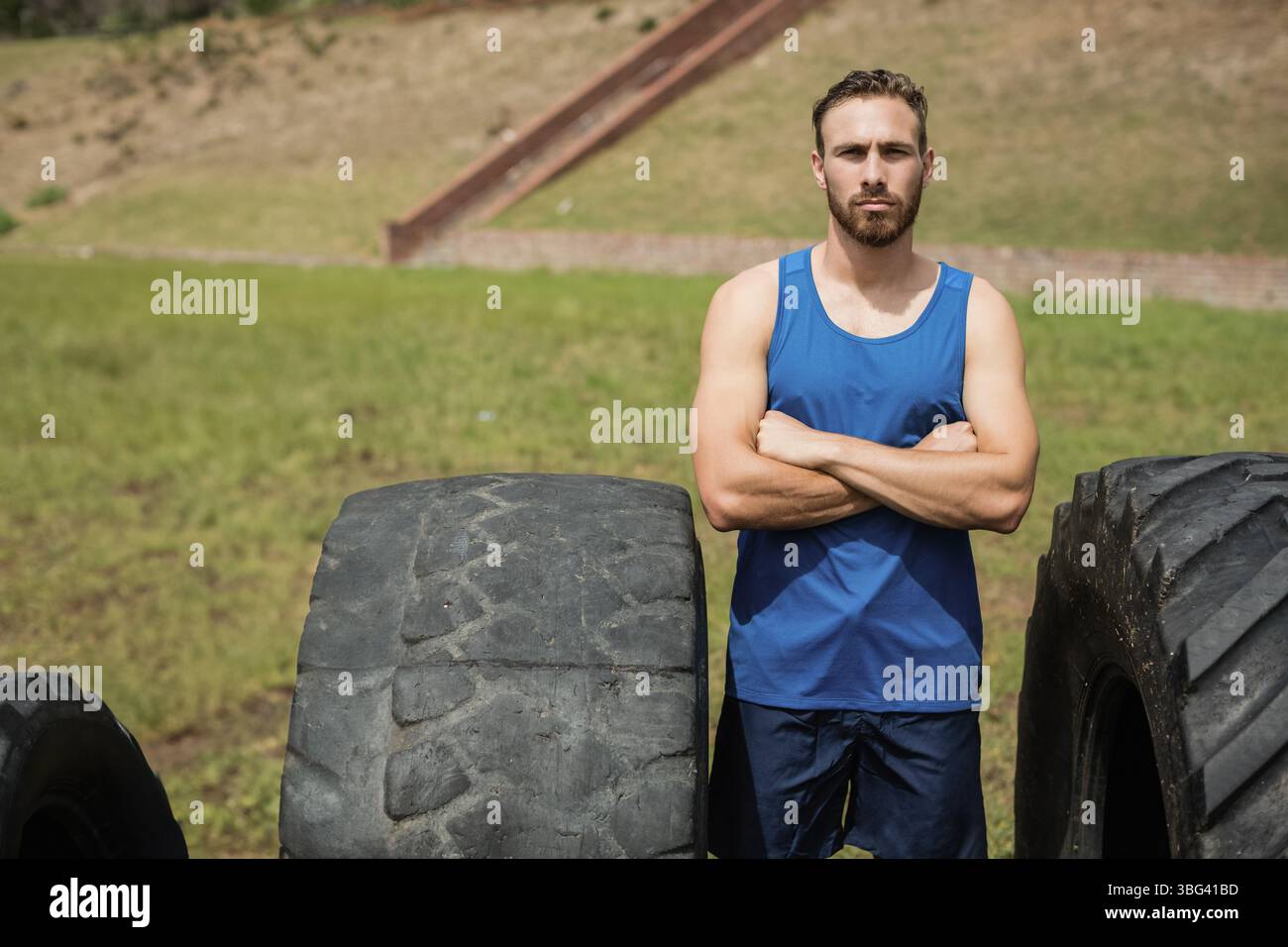 Männlicher Athlet, der auf grasbewachsenem Hang neben übergroßen Traktorreifen steht, um Krafttraining zu betreiben Stockfoto