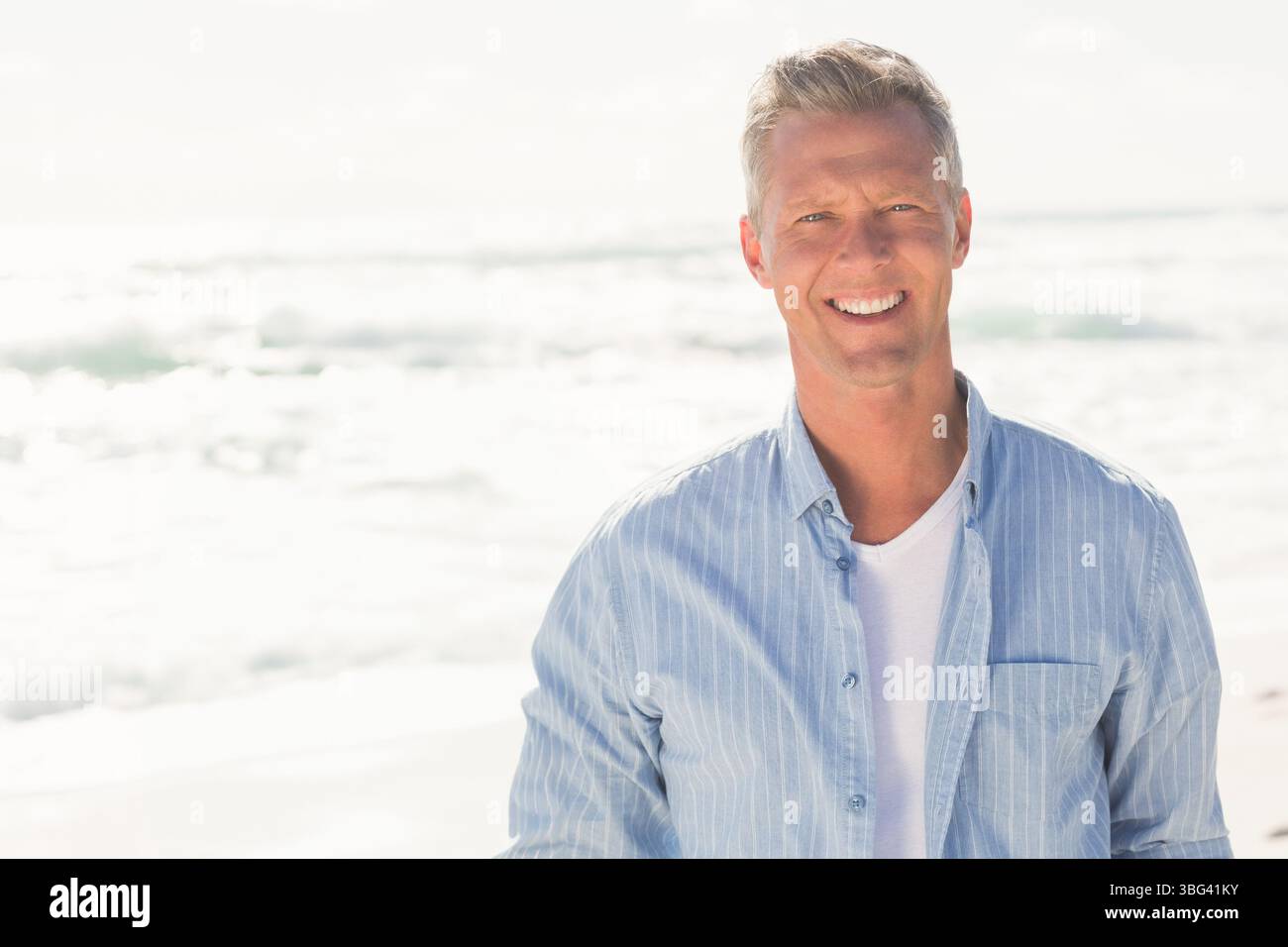 Mann lächelt, während er am Sandstrand unter Sonnenlicht steht, trägt ein blau gestreiftes Hemd, Kopierraum Stockfoto