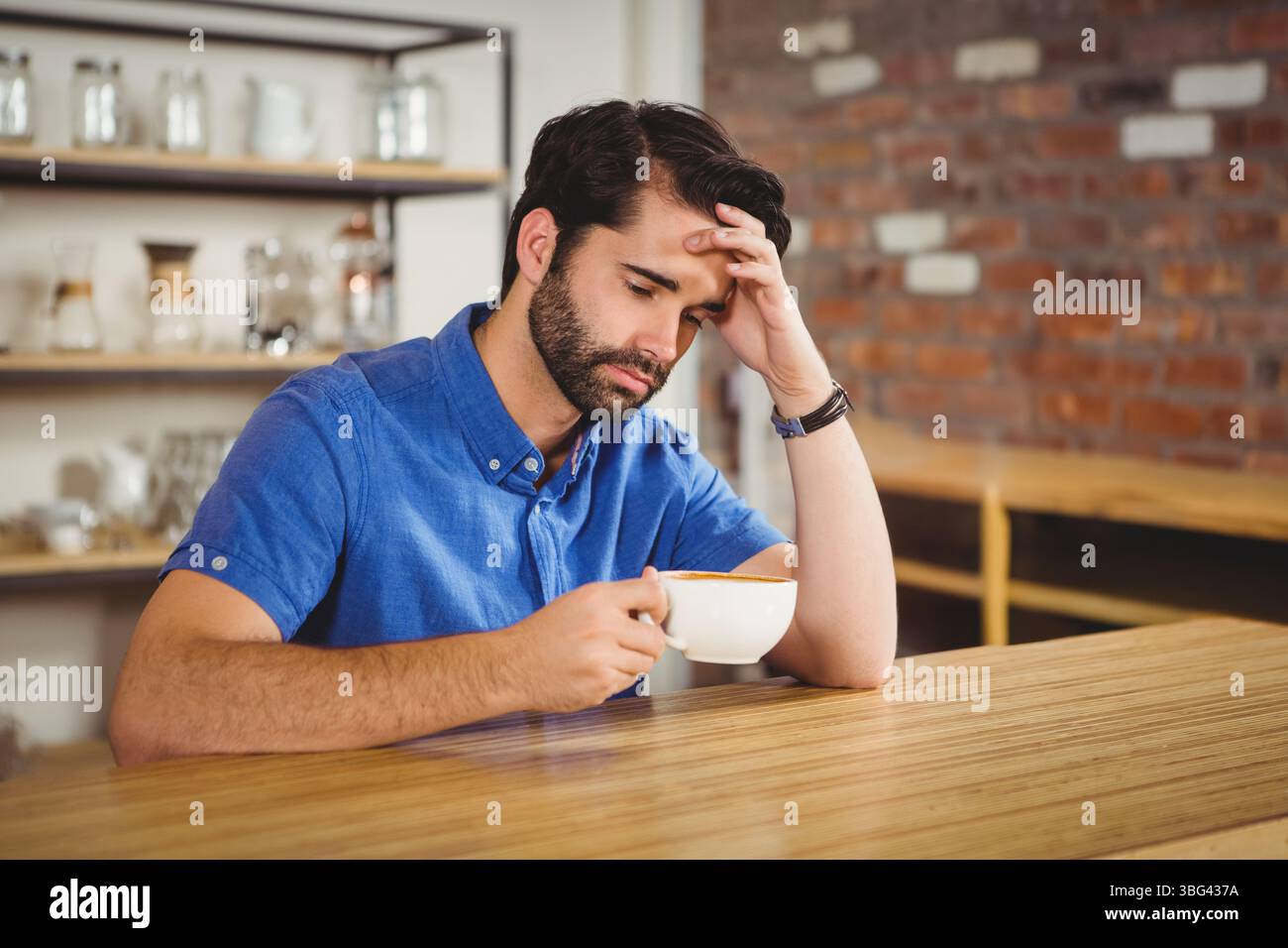 Ein einsamer Mann sitzt am Holztisch im Café und blickt auf die Kaffeetasse in der Nähe offener Regale Stockfoto
