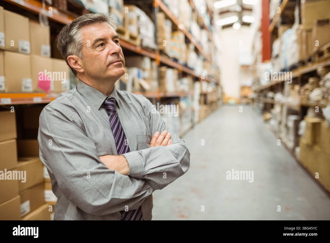 Ein stehender Mann mittleren Alters mit Hemd und Krawatte, der den Lagerbestand im Lager durch beschriftete Regale überprüft Stockfoto