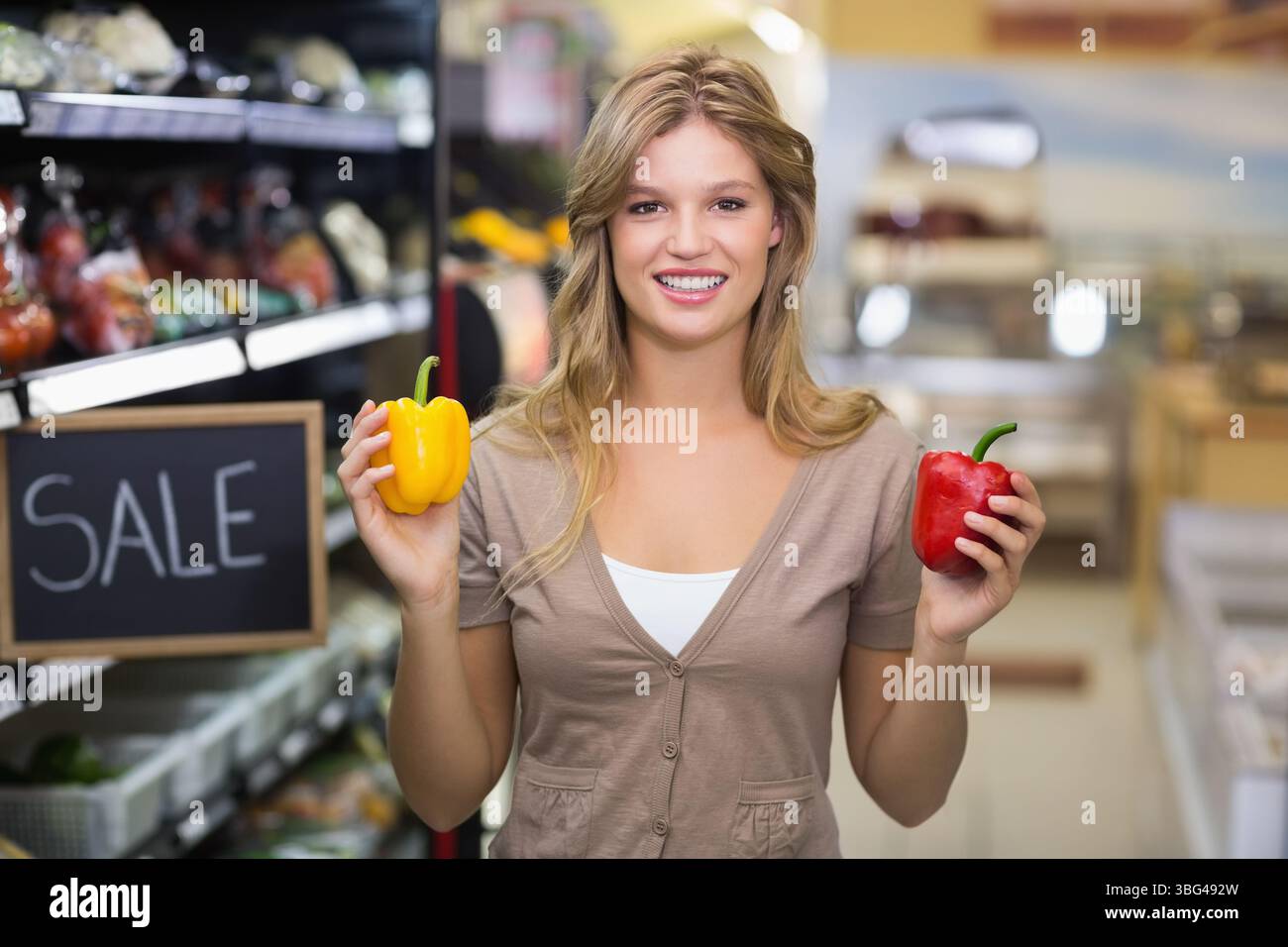 Frau in ihren Zwanzigern, die gelbe und rote Paprika im Lebensmittelgang mit Verkaufsschild hält Stockfoto