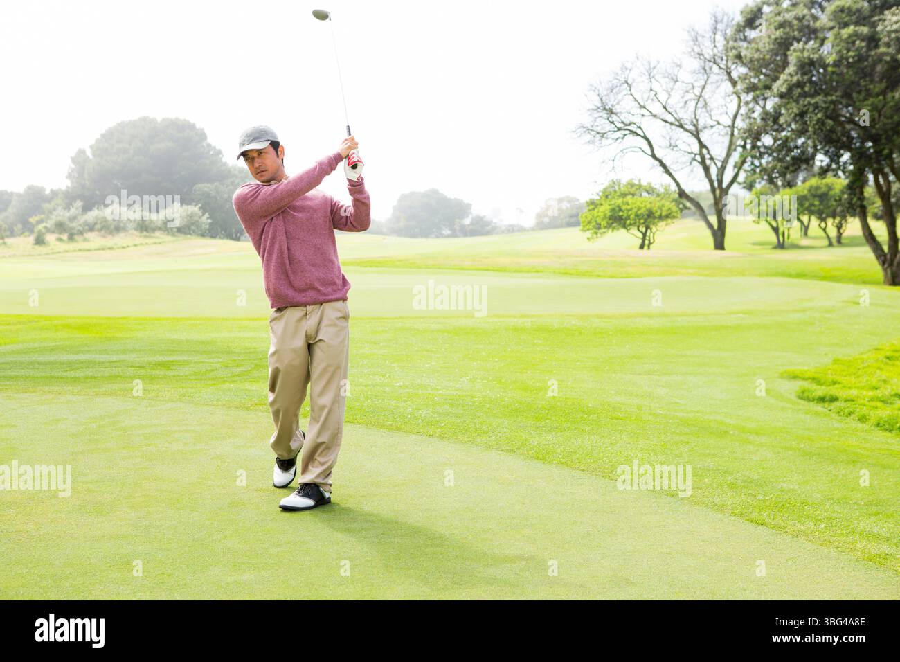Asiatischer Mann, der an sonnigen Tagen auf einem von Bäumen gesäumten Fairway auf dem Putting Green mit Golfeisen steht Stockfoto