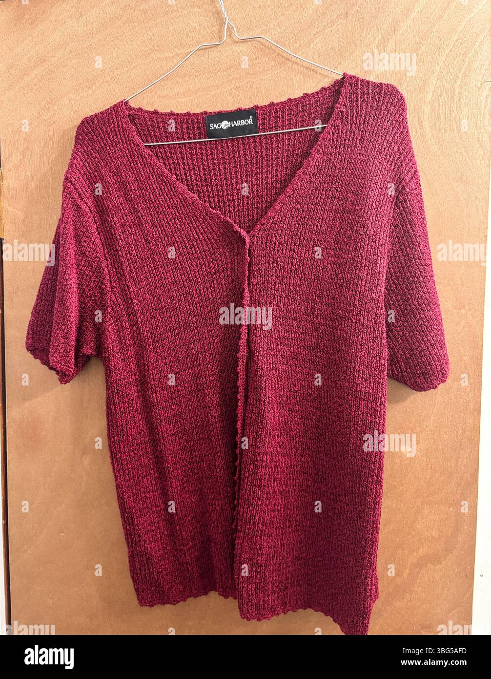 Ein strukturierter roter Sag Harbor Kurzarm-Strickcardigan hängt an einem Drahthaken vor einem warmen Holzpaneel-Hintergrund. Stockfoto