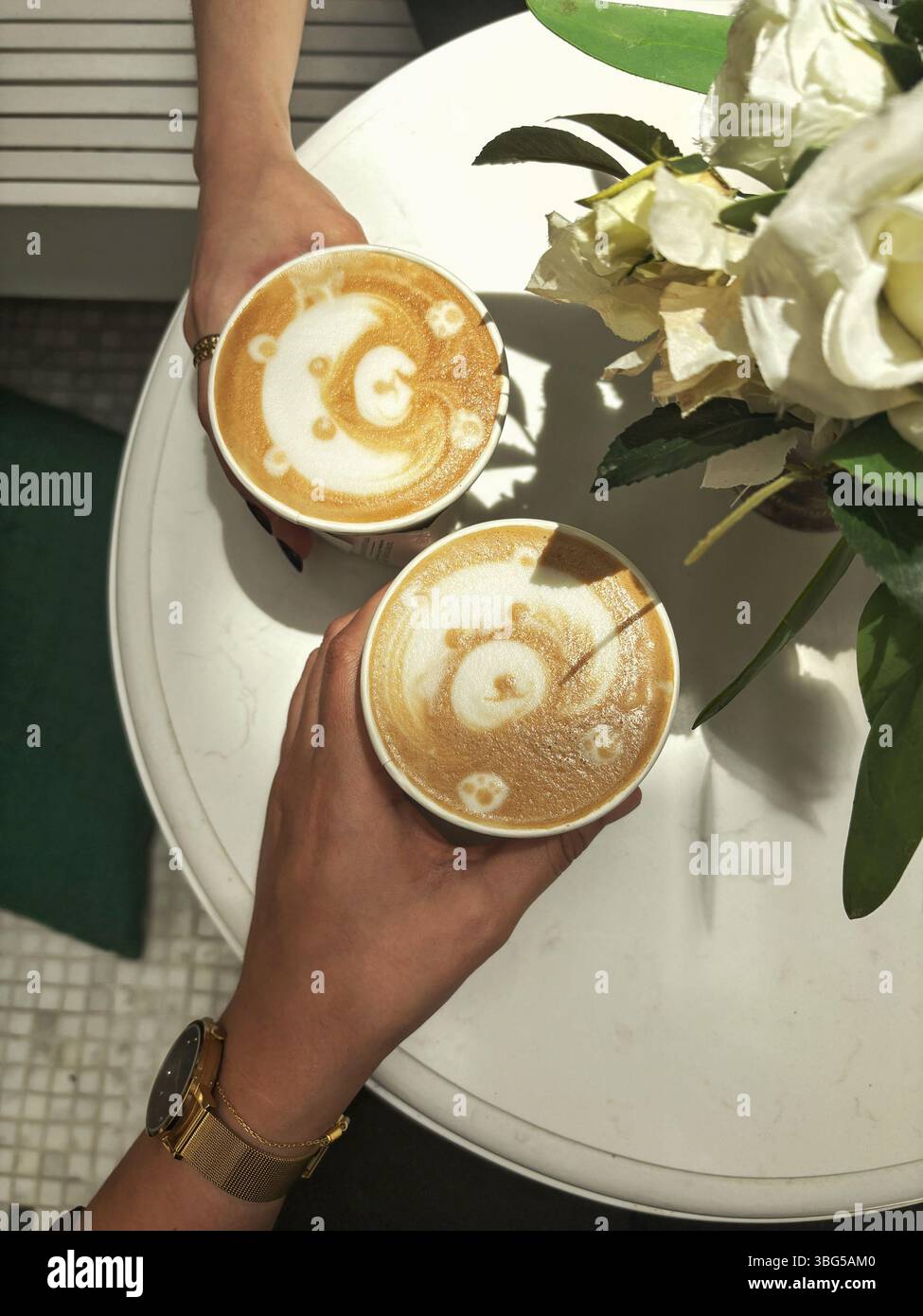 Zwei Kaffeetassen mit bärenförmigem Latte Art stehen auf einem weißen Tisch neben einem Strauß weißer Rosen. Stockfoto
