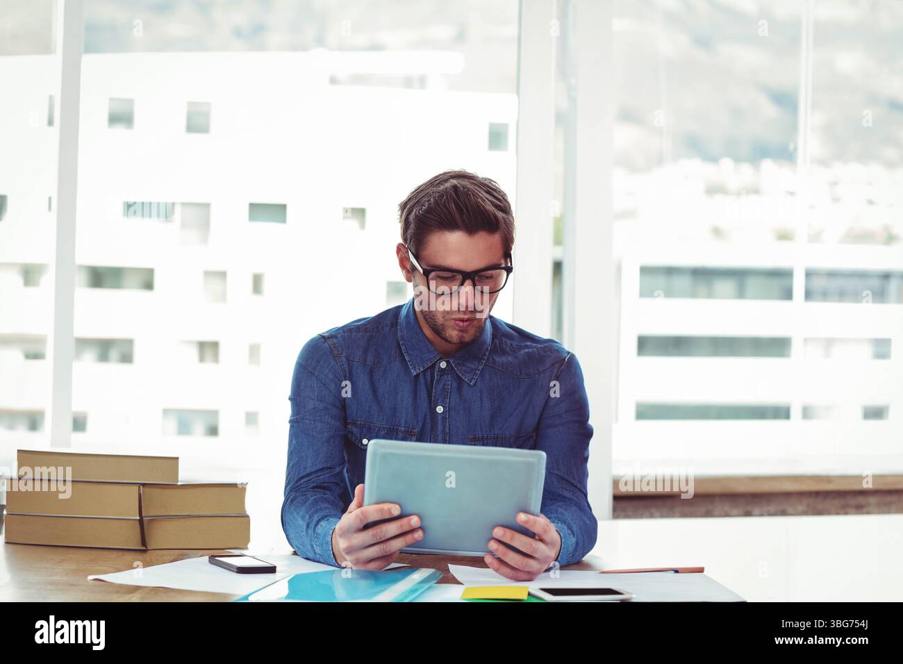 Das Tablet liegt an hohen Fenstern auf dem Schreibtisch, Bücher und Smartphones sind angeordnet, Tageslicht streamt Stockfoto