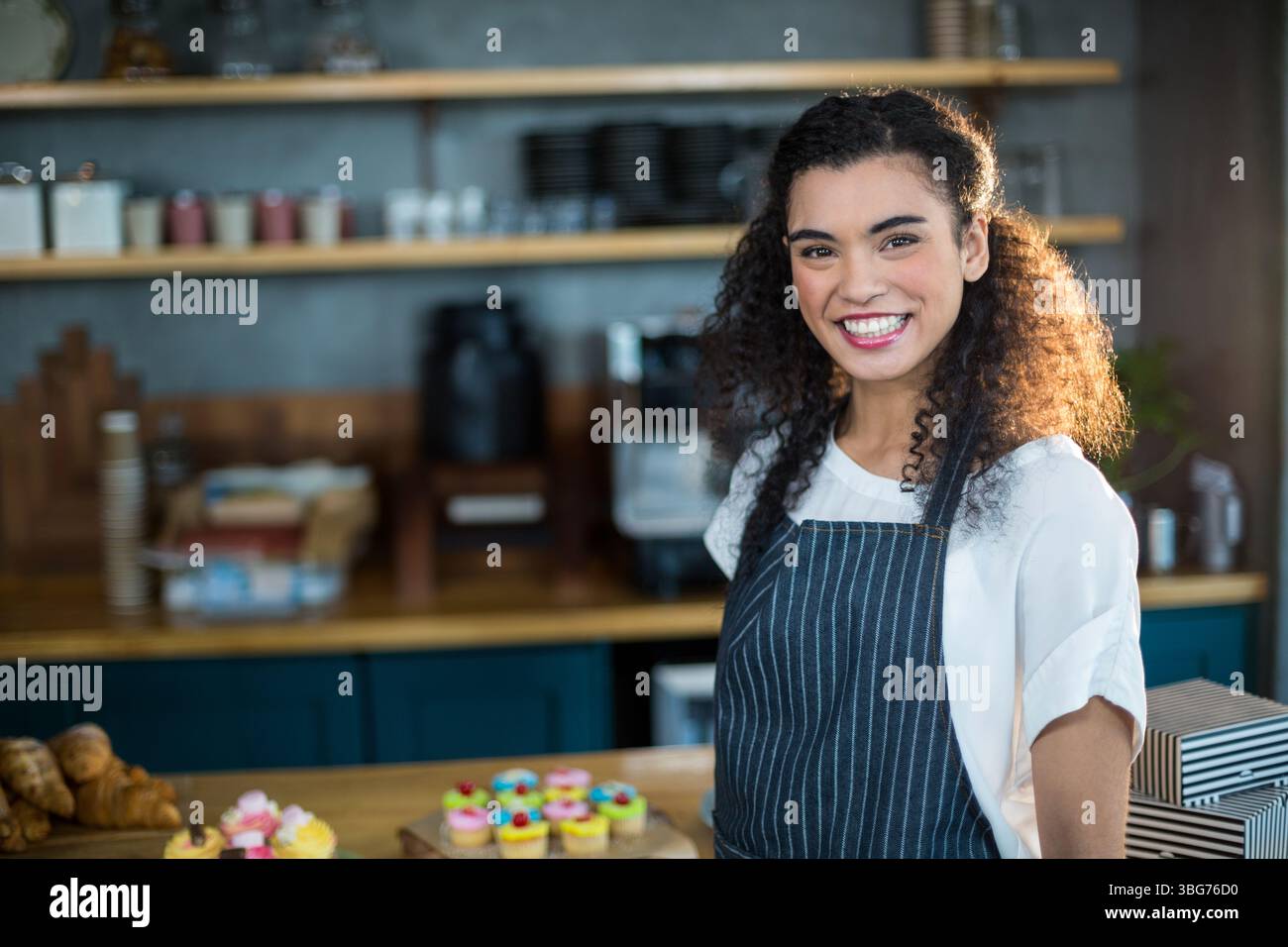 Lächelnde Afroamerikanerin serviert Cupcakes hinter der Theke im Café mit Schürze, Kopierraum Stockfoto