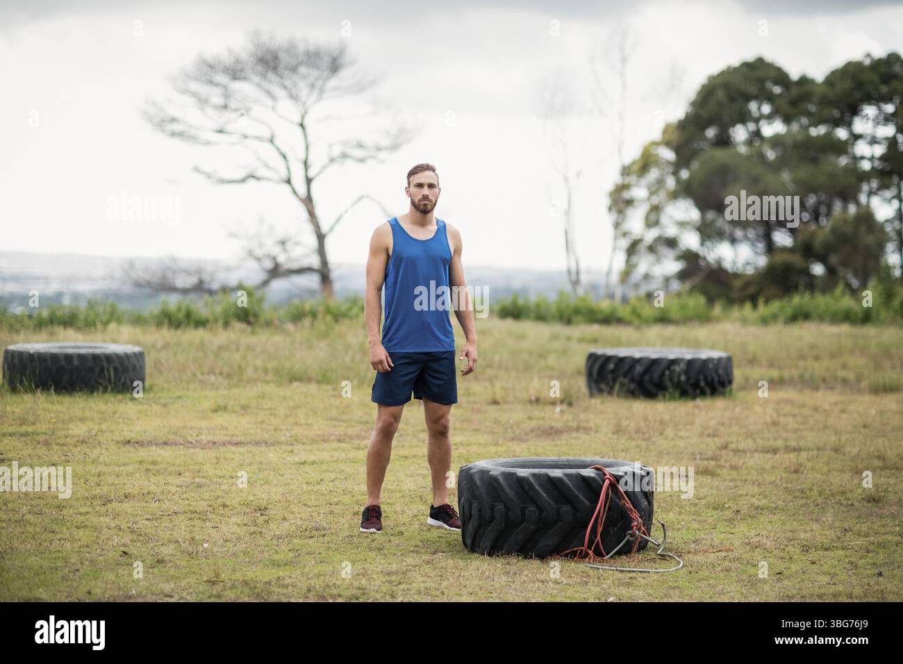 Mann mit Sportbekleidung, der auf einem grasbewachsenen Feld steht und einen mit Seilen umwickelten Traktorreifen unter bewölktem Himmel hält Stockfoto