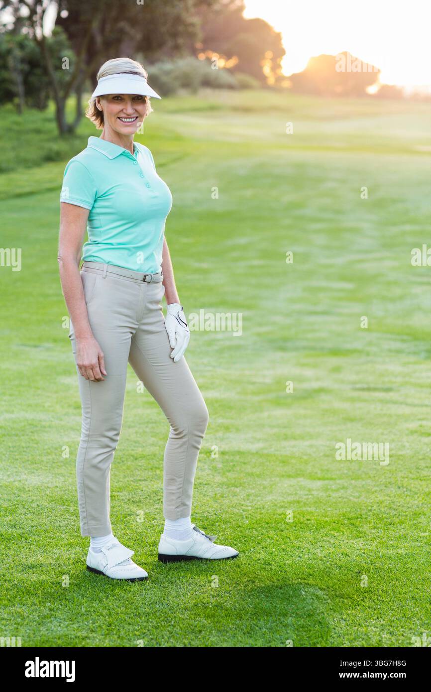 Ältere Frau, die bei Sonnenuntergang auf dem Golf Fairway steht, Visier, Golfhandschuh, Golfschuhe, Kopierraum trägt Stockfoto