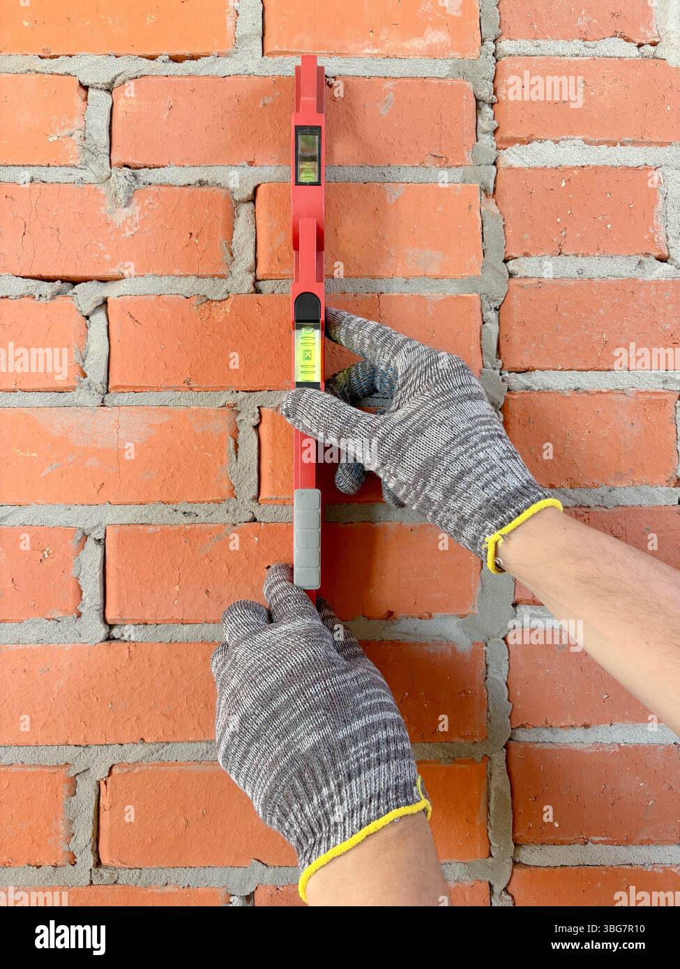 Ein Baumeister verwendet eine Luftwaage, um eine neue rote Ziegelwand in Innenräumen zu überprüfen. Bau und Renovierung von Gebäuden. Vertikales Foto mit den Händen und waagerecht auf Stockfoto
