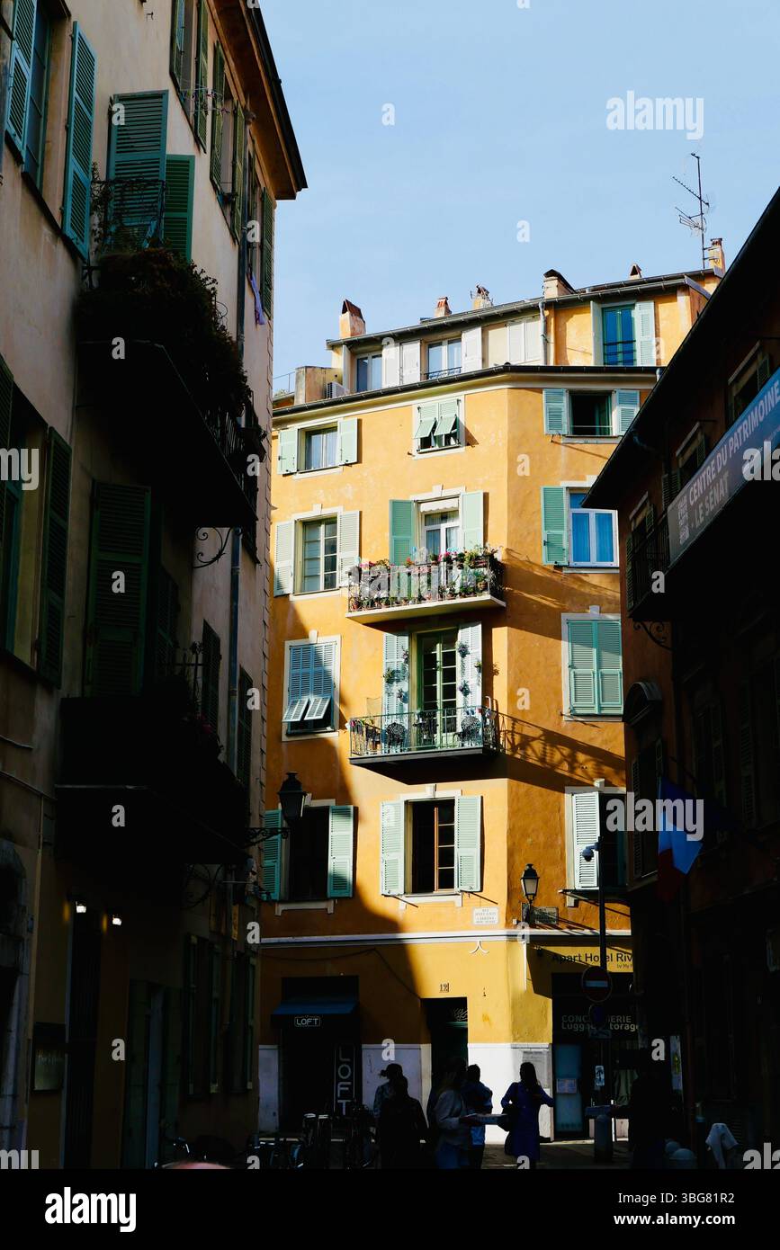 Gelbes, sonnendurchflutetes Apartmentgebäude in der Altstadt von Nizza, Frankreich, von einer schattigen Gasse mit klassischen französischen Fensterläden und Balkonen aus gesehen. Stockfoto