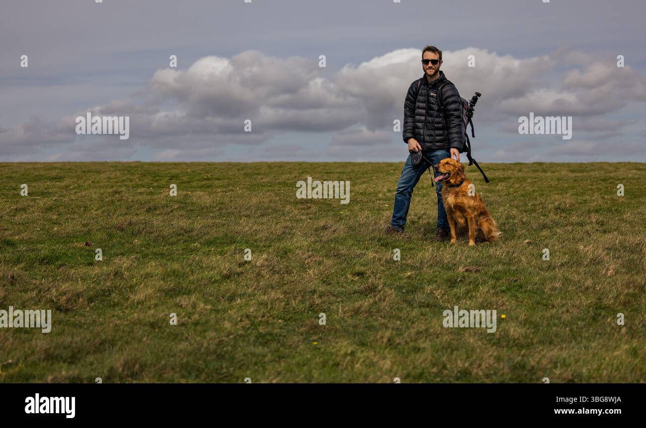Mann mit goldenem Retriever, der auf einem grasbewachsenen Hügel unter einem dramatischen bewölkten Himmel steht, Outdoor-Wanderung in der Natur, Großbritannien. Stockfoto