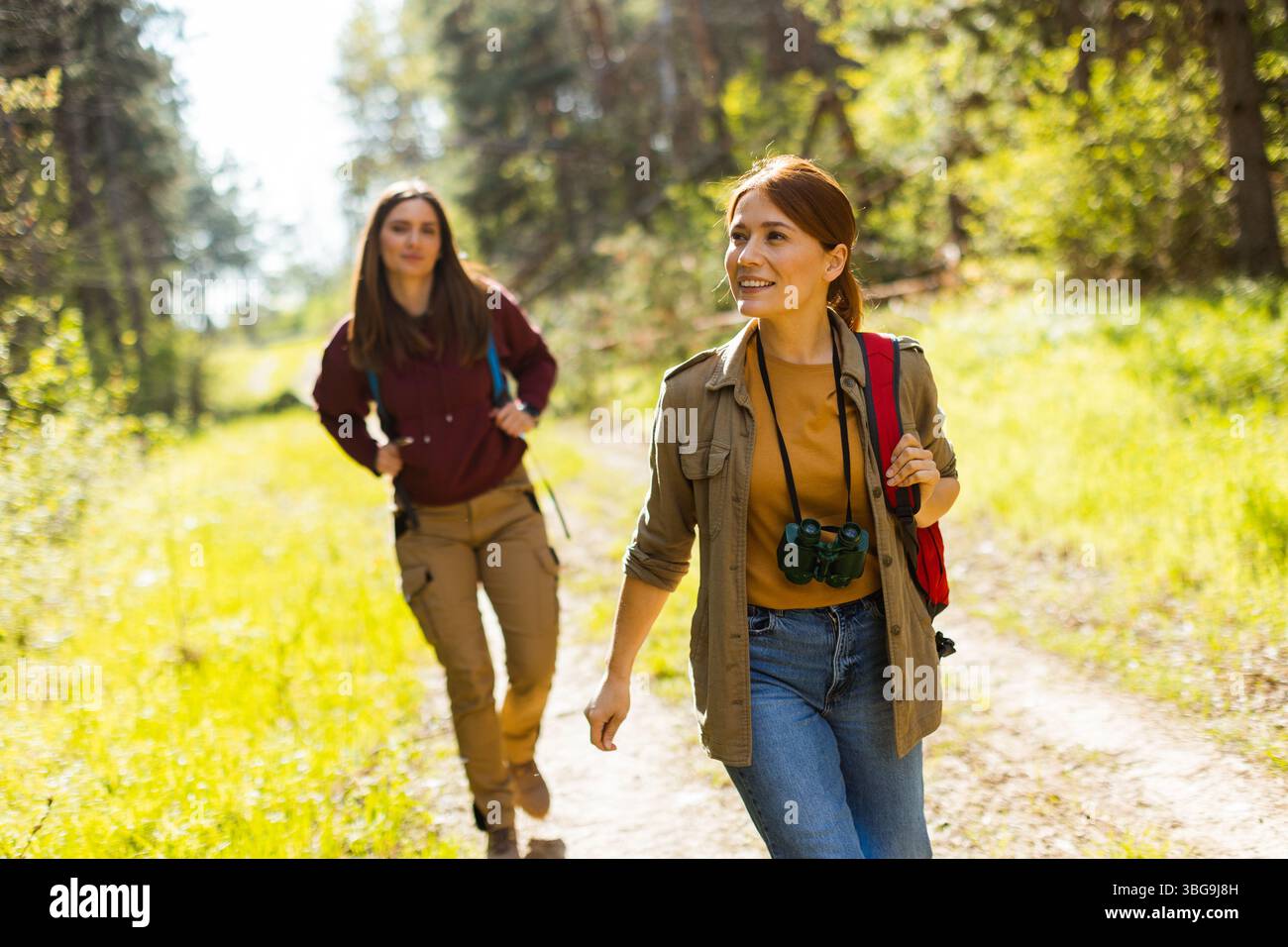 Zwei Freunde schlendern auf einem üppigen Waldweg, umgeben von Grün, teilen ein Lächeln und genießen die Wärme des sonnigen Tages. Stockfoto