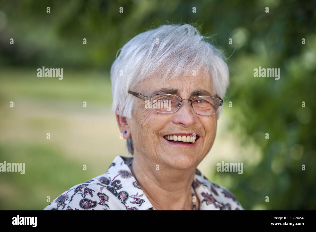 Kopfporträt einer 79-jährigen Frau mit weißem Haar vor dem verschwommenen Hintergrund eines Stadtparks, die glücklich über die Kamera lacht Stockfoto