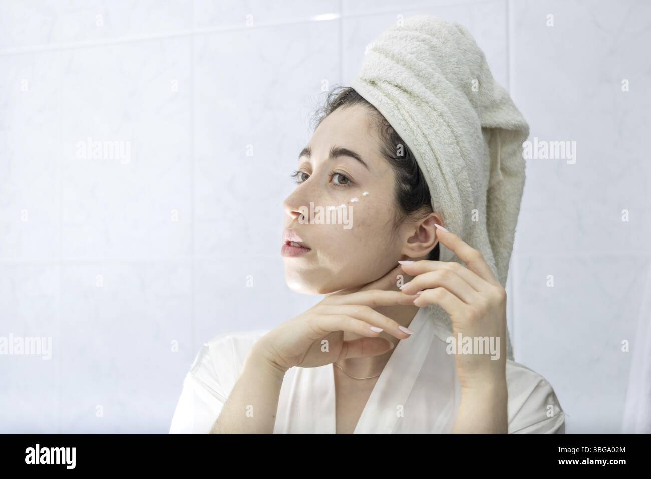 Junge Latina Frau, die in den Spiegel blickt, während sie nach einem entspannenden Bad Creme auf ihr Gesicht aufträgt.Pflege und Wellness Konzept Stockfoto