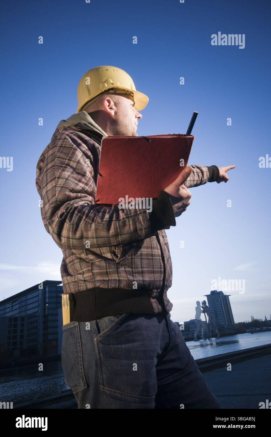 Seitliche Dreiviertelansicht eines Bauarbeiters mit gelbem Helm, karierter Jacke und rotem Losblattbinder mit dem linken Arm in den dist Stockfoto