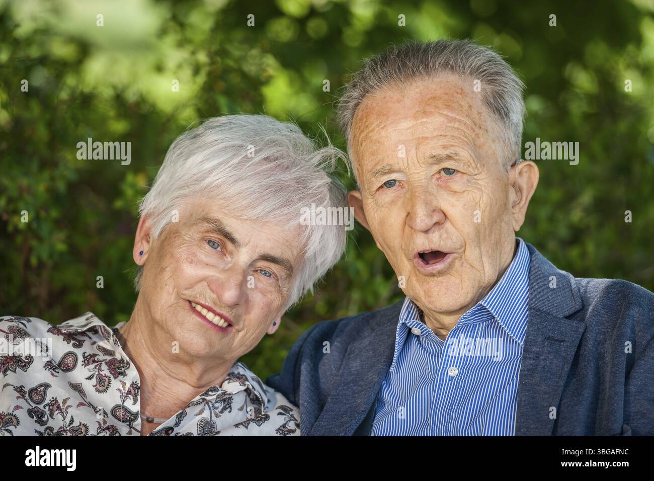 Kopf-Schulter-Porträt eines älteren Rentnerpaares vor grünen Blättern im Sommer, der fröhlich in die Kamera blickt. Mann mit offenem Mund, Talki Stockfoto