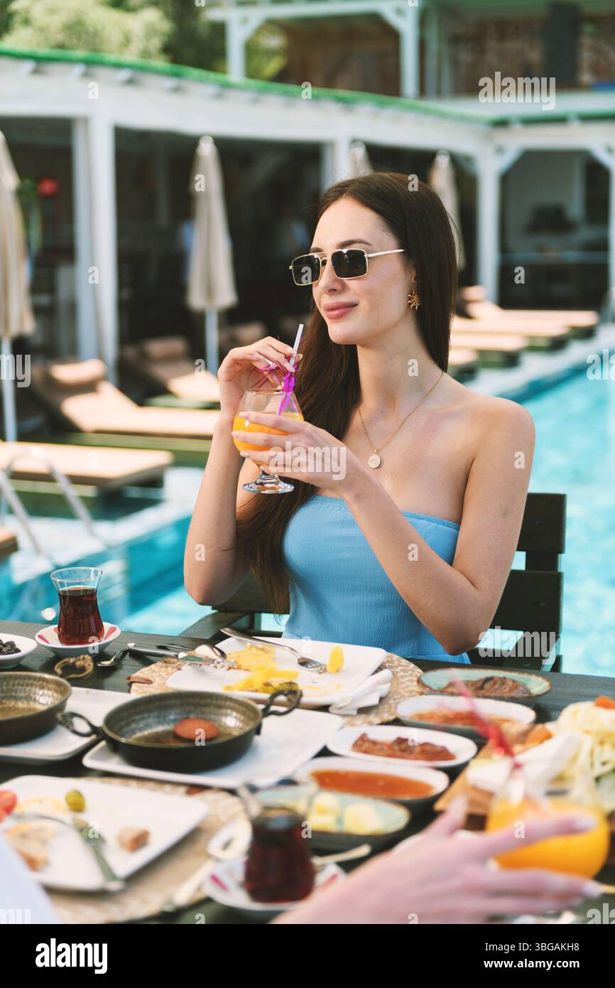 Eine brünette Frau aus den 20er Jahren serviert ein üppiges Frühstück mit Orangensaft in der Hand und bietet den Hotelpool im Hintergrund. Stockfoto