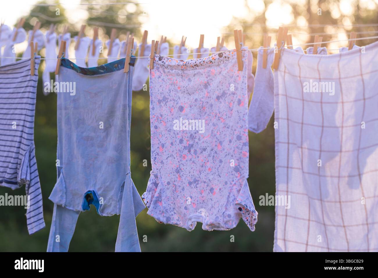 Bunte Familienkleidung, die an einem sonnigen Nachmittag im Freien trocknet, ruhige ländliche Landschaft und wenig Sonnenlicht, das durch die Bäume strahlt, vermitteln ein warmes Zuhause Stockfoto