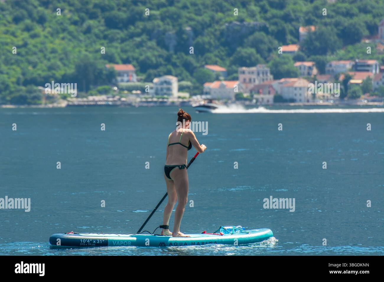 Junge Frau Standup Paddleboarding (SUP) in Kotor Bay, Montenegro mit traditionellem montenegrinischen Dorf im Hintergrund. Stockfoto