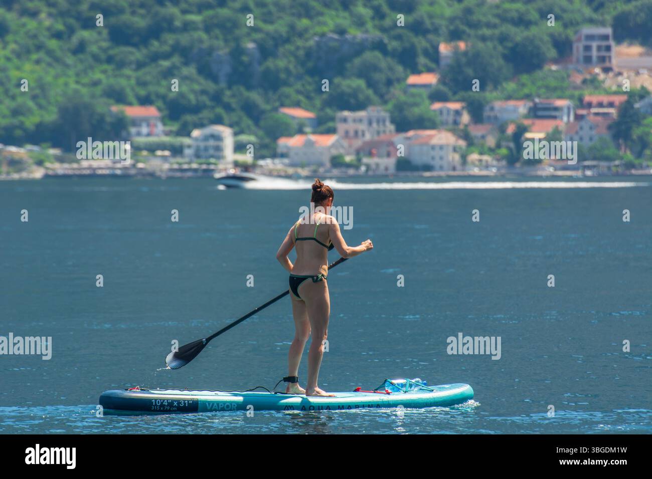 Junge Frau Standup Paddleboarding (SUP) in Kotor Bay, Montenegro mit traditionellem montenegrinischen Dorf im Hintergrund. Stockfoto